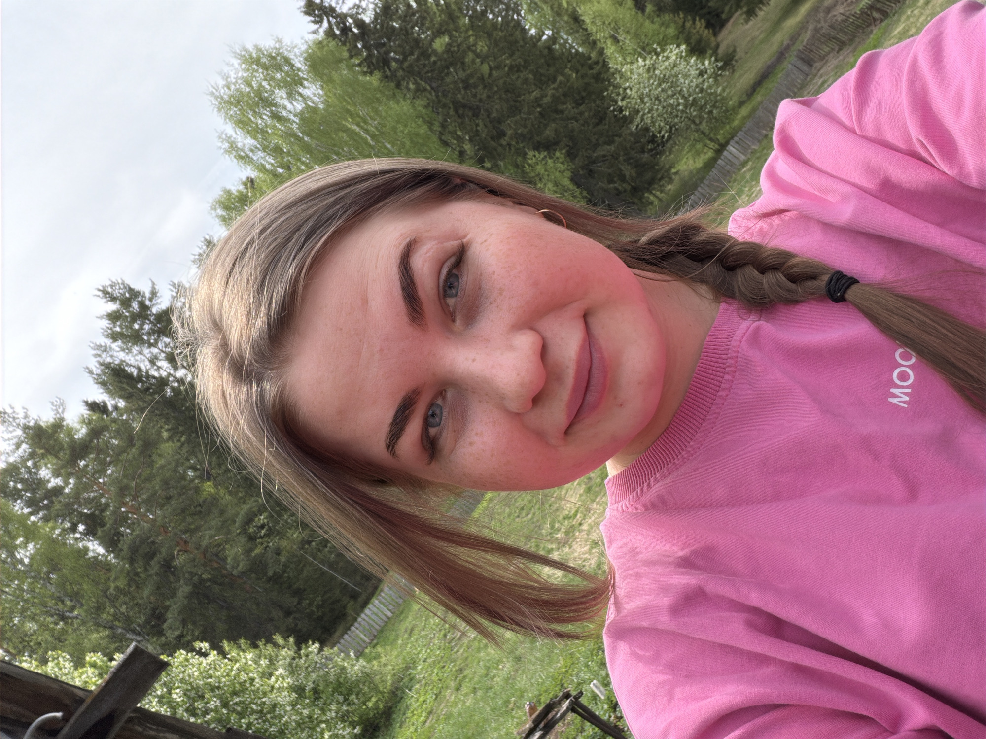 The image shows a young woman with light brown hair, partly braided on one side and secured with a black hair tie. She is wearing a pink sweatshirt with the word "MOON" partially visible. She has fair skin with freckles and blue eyes. The background consists of trees and a grassy area, suggesting an outdoor setting. The photo appears to have been taken during the daytime under cloudy skies. The location is Уральское, Russia.