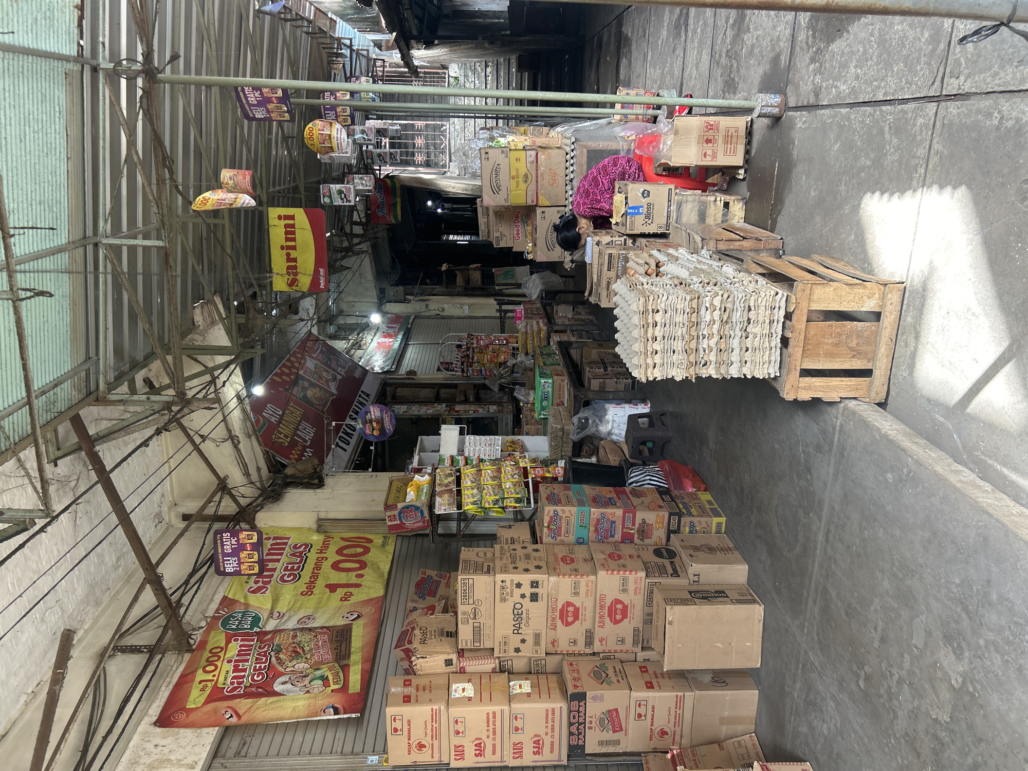 The image shows a street-level market stall in Denpasar, Indonesia, likely a small grocery or warung. The stall is covered by a corrugated metal roof supported by metal poles. Various boxes of packaged goods, including what appear to be instant noodles, sauces, and detergents, are stacked high on either side of the walkway. Several promotional banners for "Sarimi" instant noodles are visible, one prominently featuring a price of Rp 1,000 and another offering a "Buy 2 Get 1 Free" deal. A woman wearing a pink patterned dress is seated on a red stool, seemingly organizing or attending to the goods. Stacks of egg cartons are piled on wooden crates in the foreground. The walkway is paved with concrete and shows signs of wear and tear. The overall scene suggests a bustling, everyday commercial environment.