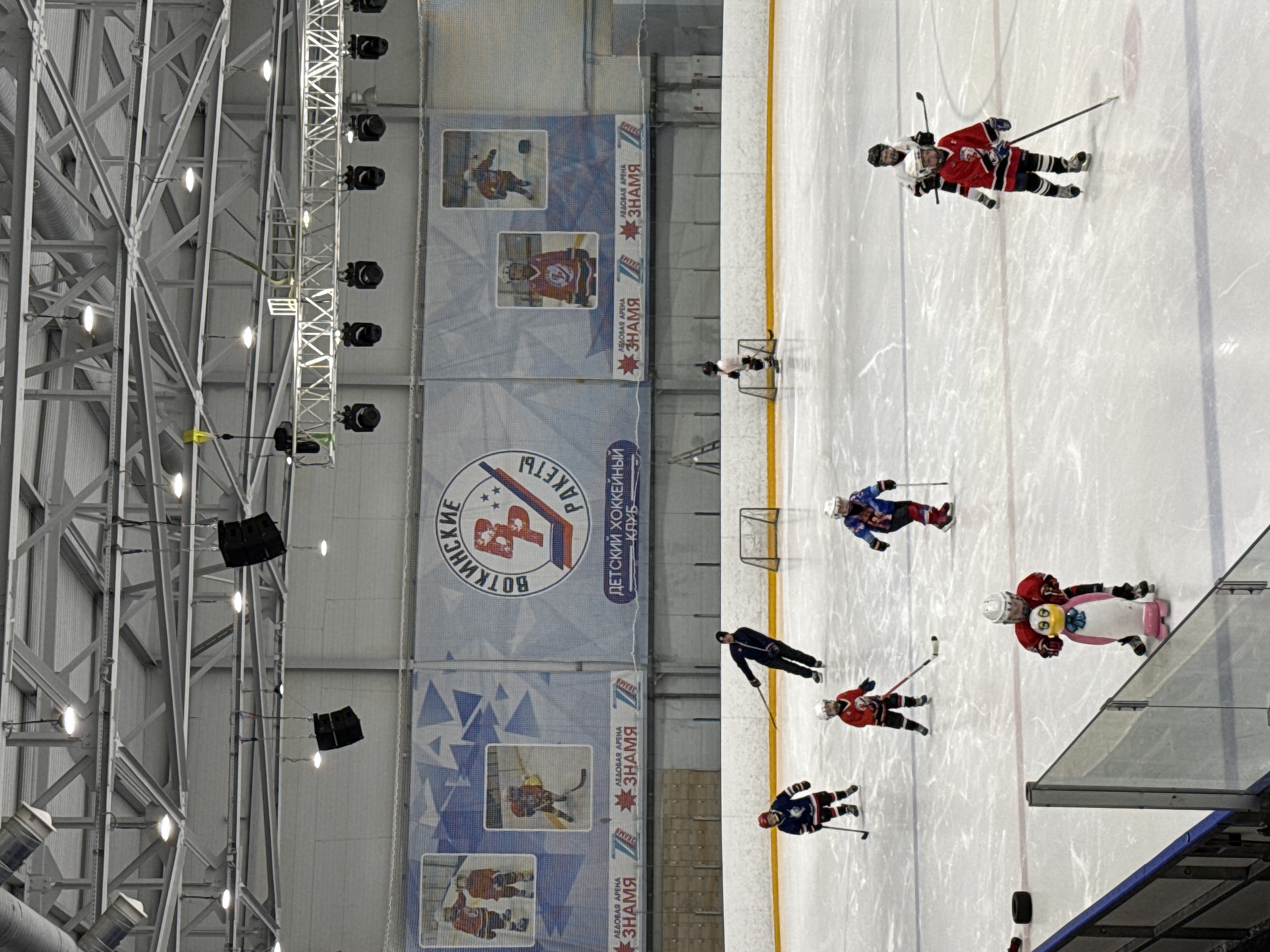The image depicts a hockey game taking place indoors at the Ice Arena Znamya, located in Votkinsk, Russia. Several young hockey players wearing different colored uniforms are skating on the ice. A coach is also visible on the ice. Banners with team logos and arena branding are displayed on the wall. Structural support beams and lighting fixtures are visible on the ceiling.