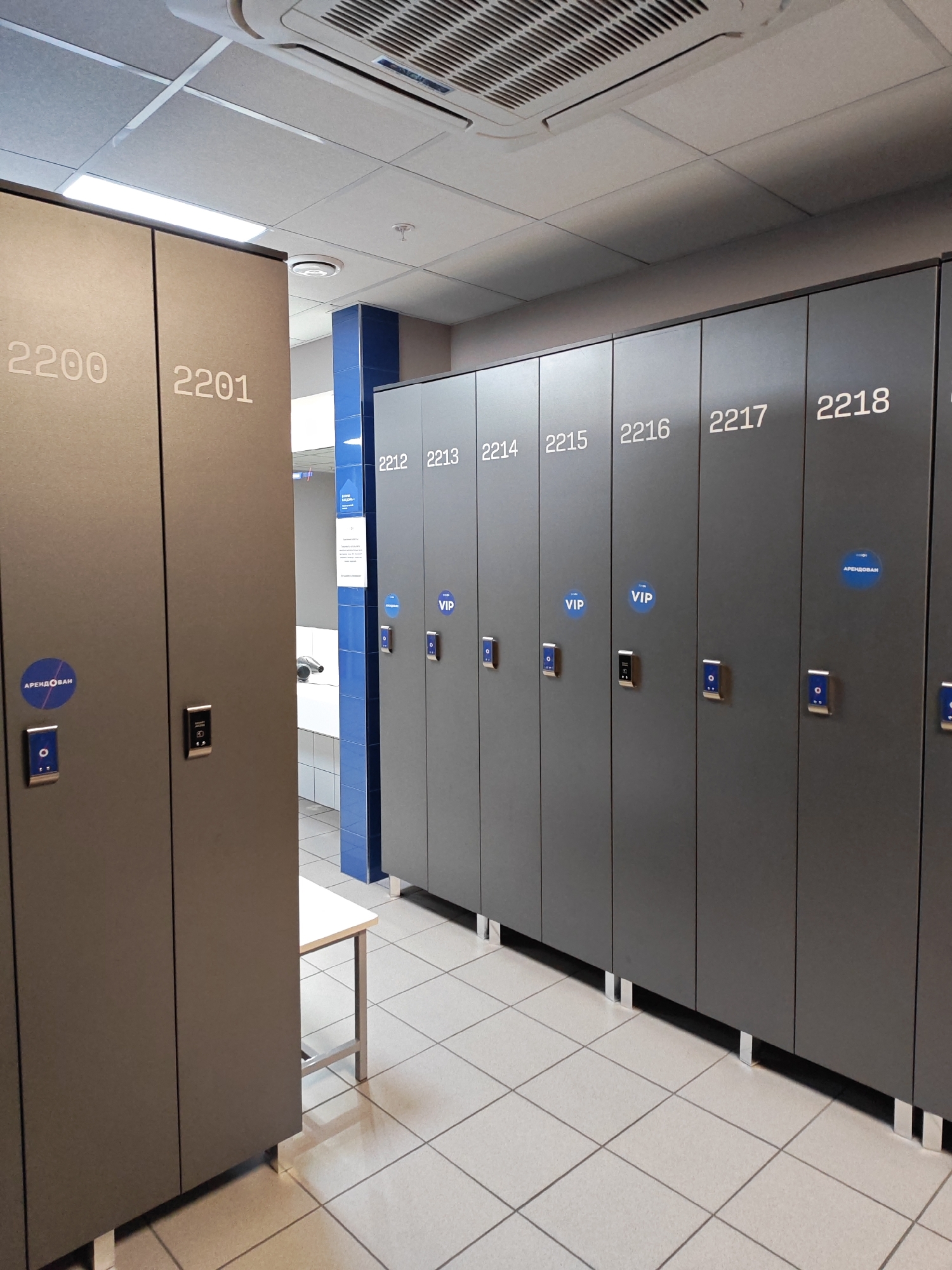 This media file depicts a row of grey lockers in what appears to be a gym or spa changing room. The lockers are numbered sequentially, starting from 2200 and 2201 on the left, and continuing with 2212, 2213, 2214, 2215, 2216, 2217, and 2218 to the right. Some lockers are marked with blue circular stickers. One sticker on locker 2200 reads "АРЕНДОВАН" (rented) in white text. Another sticker on locker 2218 also has the same text. Two lockers, numbered 2215 and 2216, have "VIP" printed on blue circular stickers.
Each visible locker door features an electronic lock mechanism. The lockers are situated in a brightly lit indoor environment, with a tiled floor and a white ceiling featuring fluorescent lighting and an air conditioning unit. A blue tiled wall is visible behind the lockers on the right side of the image. A small white bench with metal legs is positioned between locker 2201 and the row of numbered lockers, suggesting a seating area for changing. The overall impression is one of order and functionality, characteristic of a public facility. No people are visible in the image. The lighting suggests it is daytime, and there are no weather cues. The presence of locker numbers and facility-related stickers indicate a functional space for patrons.