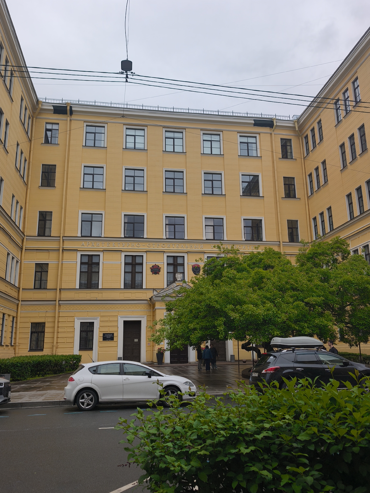 Here is a detailed description of the image:
The image captures an urban scene in Saint Petersburg, Russia. It features a yellow building, identified by the inscription "ARCHITECTURNO-STROITELNY UNIVERSITET" (Architectural and Construction University) above its entrance. This indicates the location is likely the Saint Petersburg State University of Architecture and Civil Engineering.
The building is a multi-story structure with a symmetrical facade. It has several windows with black frames and a cornice along the top. The entrance is centered and features a small decorative pediment. To the right of the entrance are two trees with green leaves.
In front of the building, there are two cars parked: a white hatchback on the left and a black SUV with a roof box on the right. Between them, near the building's entrance, several people are present, including two standing in front of the door. A green bush occupies the foreground, partially obscuring the cars.
Above the building, there are several black power cables, obscuring the sky. The scene is captured on a cloudy day, with the pavement appearing wet, suggesting recent rain.