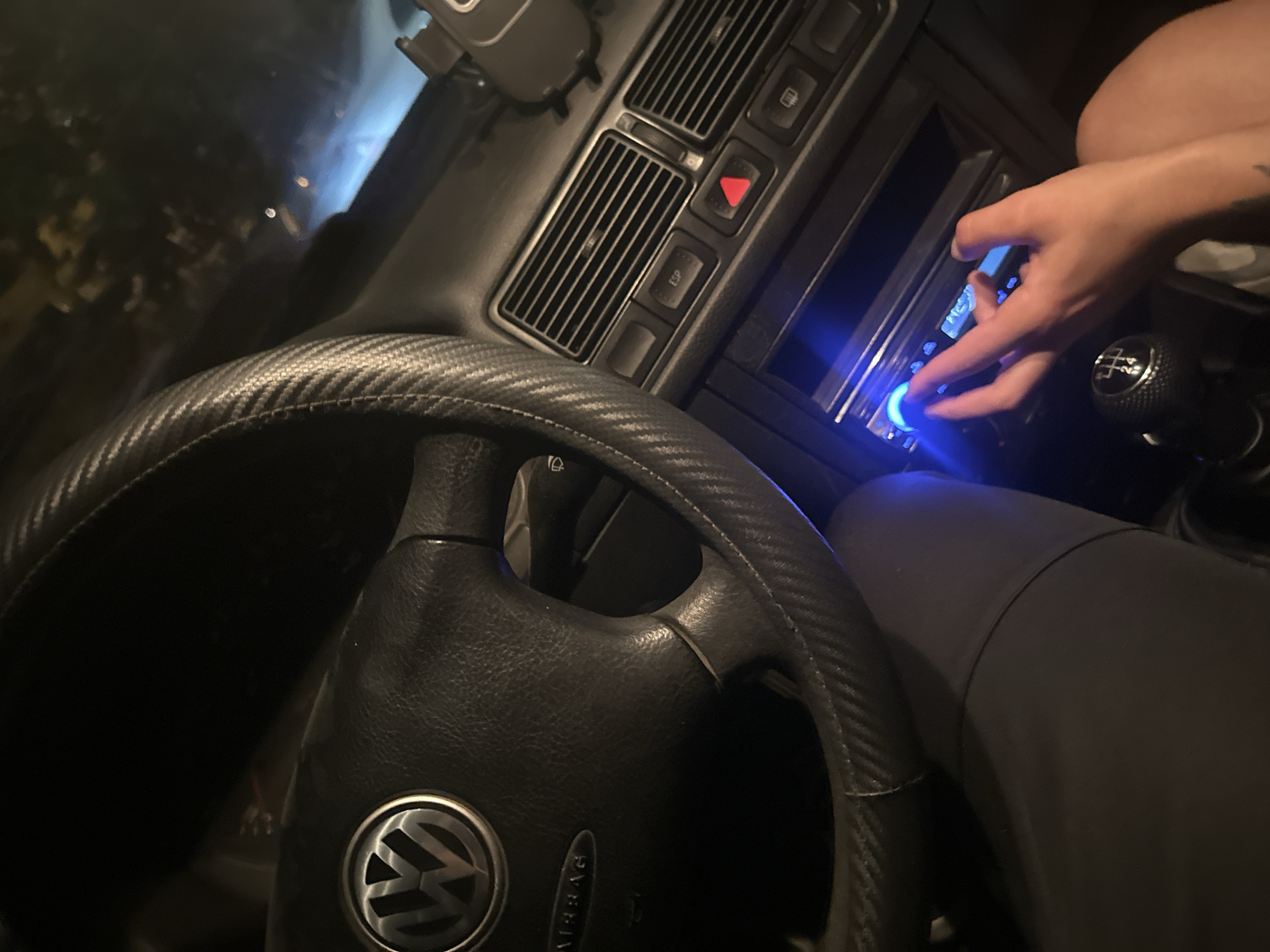 The interior of a Volkswagen vehicle is visible. The steering wheel, adorned with a carbon fiber-like finish, features the Volkswagen logo. A hand is manipulating a car stereo system with blue illumination. A gear shift is located to the right. The environment suggests the inside of a car in an unidentifiable place. This image is not connected with the city of Plovdiv, Bulgaria, and does not contain any visual indicators to suggest its location.