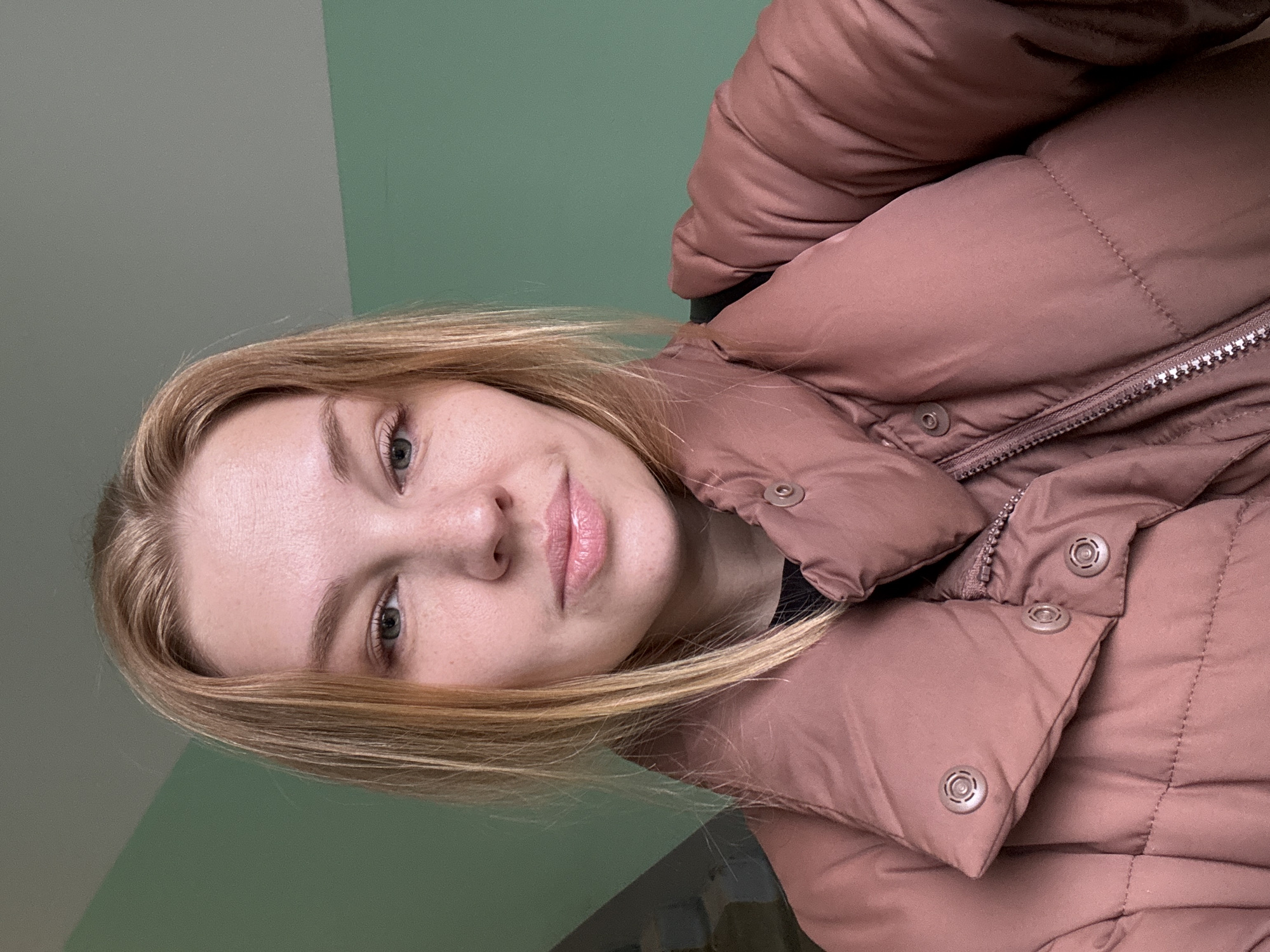A young woman with blonde hair and fair skin is facing the camera, wearing a rose-colored puffer jacket. The background is a two-toned green wall. The location cannot be determined as Rusanovo, Russia, from the image alone.