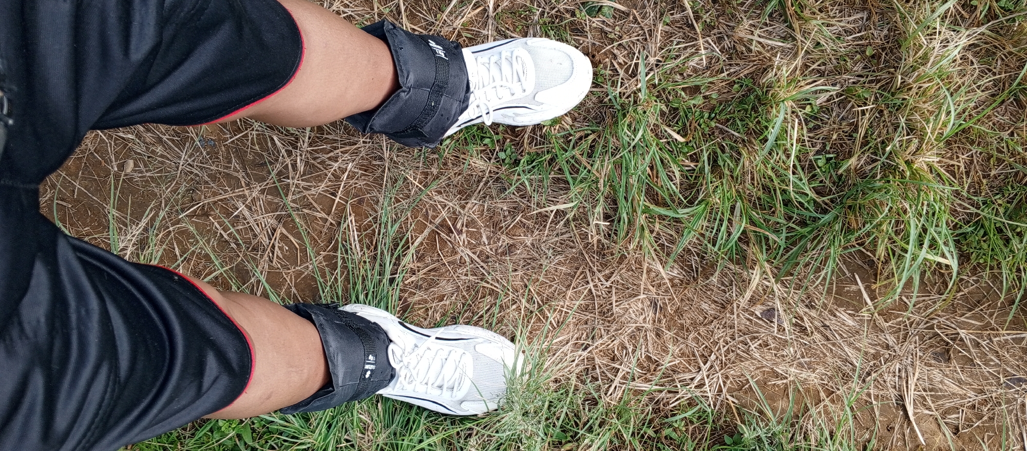 An overhead perspective captures the lower extremities of an individual standing on an outdoor surface in Weddagala, Sri Lanka. The subject wears black athletic shorts with a thin red trim visible at the leg openings. Black fabric ankle weights are secured around both ankles, positioned above white athletic shoes with white laces. Visible text on the left ankle weight indicates "1kg". The ground consists of uneven brown soil interspersed with dry, light brown grass and scattered patches of green grass. The vegetation is sparse in some areas, revealing more exposed earth.