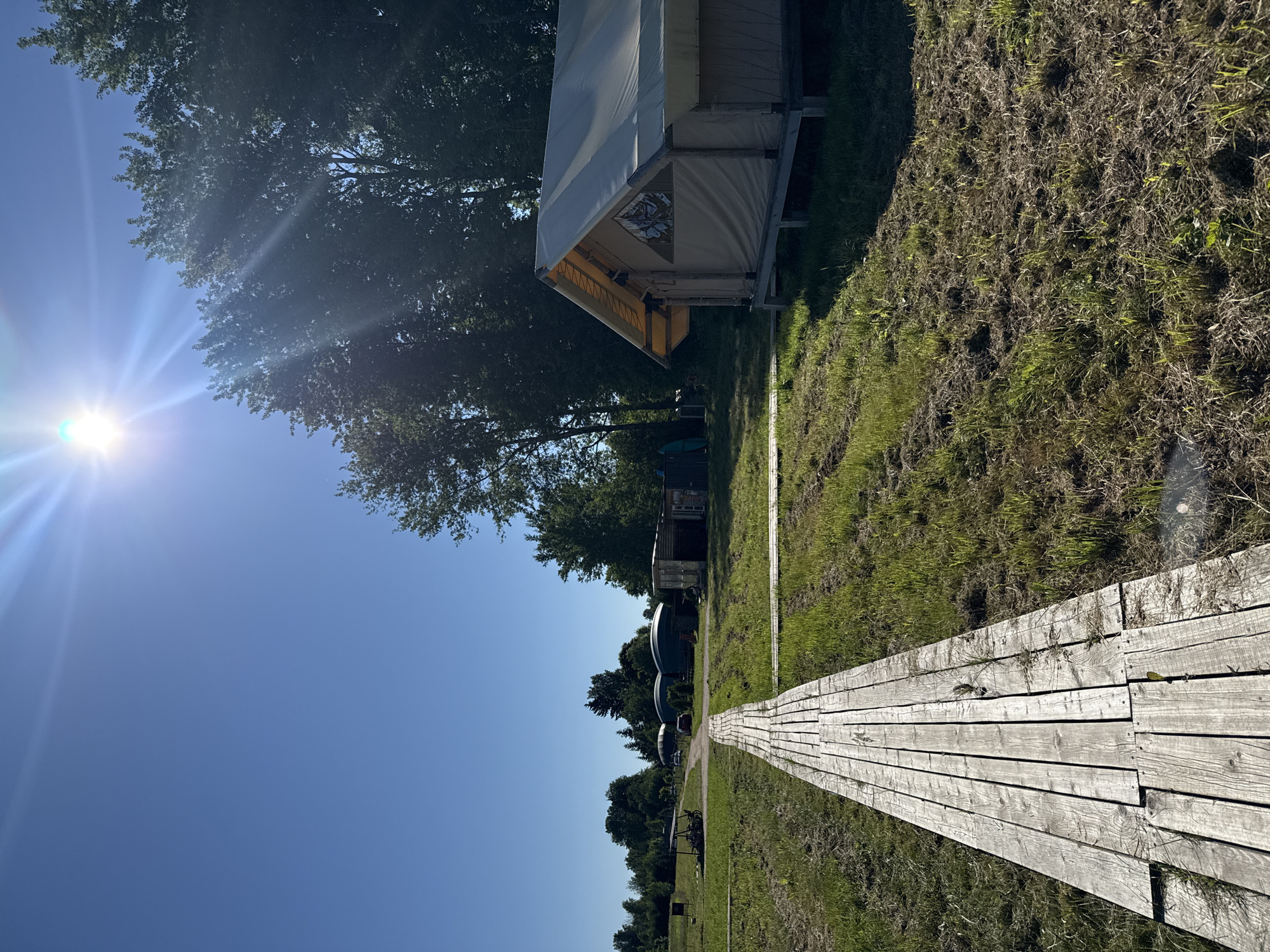 A wooden boardwalk extends into the foreground, leading towards a glamping tent on the right side of the frame. The tent is elevated on stilts and has a light-colored canvas roof and walls. In the background, there are several dome-shaped structures, some smaller sheds, a vehicle, and trees under a clear blue sky with bright sunlight and lens flare. The ground is covered in dry grass and some patchy green grass. This scene is located in the city of Zamosh'e (Zaklinskoe S/P), Russia.