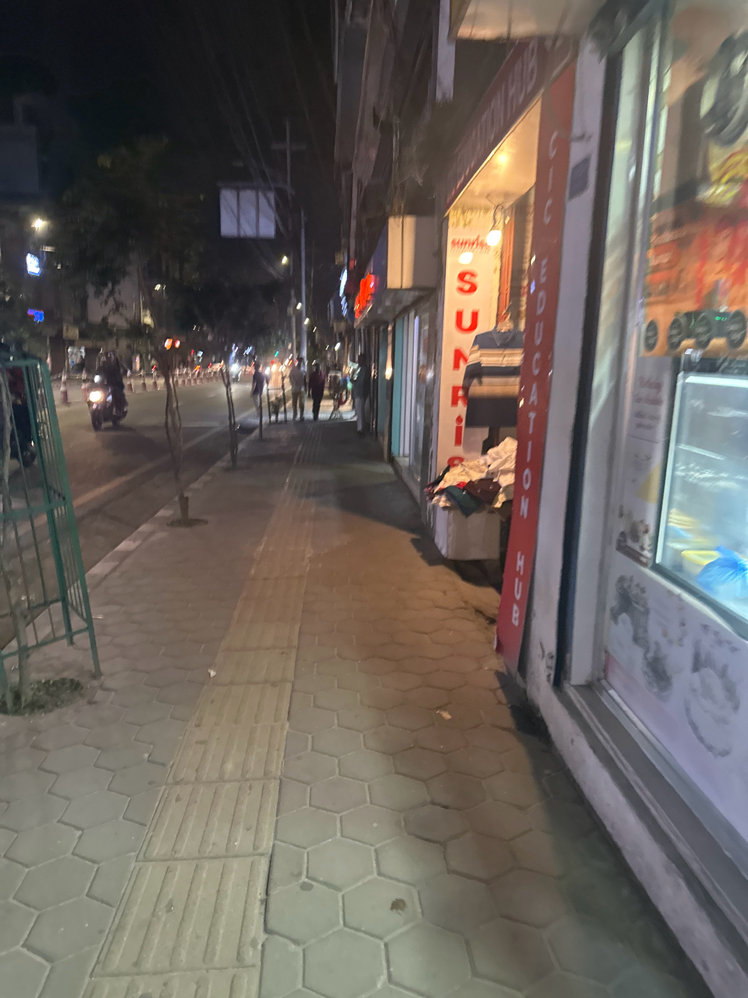 The image depicts a street scene at night in Kathmandu Metropolitan City, Nepal. The perspective is from a sidewalk looking towards a road and shopfronts. On the right, a shop window displays various items, and a refrigerator unit is visible. A red sign with white lettering, partially obscured, reads "CIC EDUCATION HUB". To the left of the shop, a clothing rack displays a striped shirt. Further down the street, several people are walking, and a motorcycle is being ridden on the road. Traffic cones are lined up along the side of the road. The sidewalk is paved with hexagonal tiles and has a tactile paving strip. Small trees are planted along the sidewalk. The overall lighting suggests it is nighttime, with streetlights and shop lights illuminating the area.