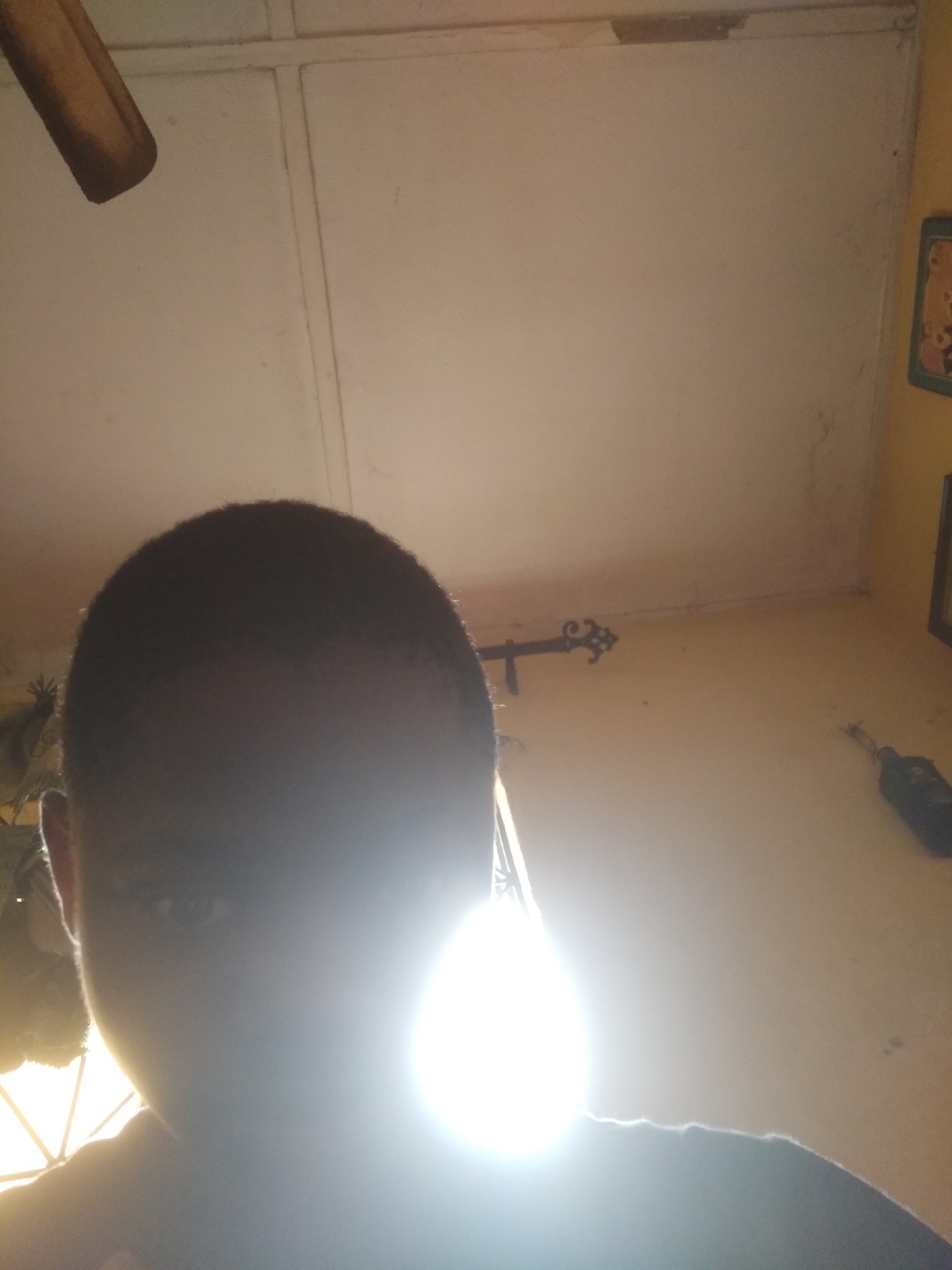 The image is a low-angle selfie taken indoors, primarily illuminated by an extremely bright light source in the lower-middle foreground. A person's head and upper body are visible in partial silhouette, with the top of their dark, short-haired head prominent in the upper-middle section of the frame. The left side of the person's face, including an eye and ear, is faintly discernible through the shadows created by the intense light, which causes significant glare and overexposure.
The background features a light-colored ceiling with visible panel lines or seams dominating the upper portion of the image. A brown, possibly wooden, beam or structural element is partially visible in the upper-left corner. Below the ceiling and to the right, a light yellow or orange-painted wall is present. Several objects are mounted on or resting against this wall:
- In the upper-right corner, a green-framed picture depicting a cartoonish character is partially visible.
- Below that, another dark-framed item is partially seen on the right edge.
- Towards the mid-right, an ornate, dark metal fixture, resembling a curtain rod finial or decorative hook, is attached to the wall.
- Further down on the wall, a small, dark object, possibly a bottle or container, is lying horizontally.
The strong direct light source heavily impacts the visibility of details and colors, casting much of the scene into shadow or overexposure. The setting appears to be a casual indoor room, possibly a home. No visible text is present in the image.