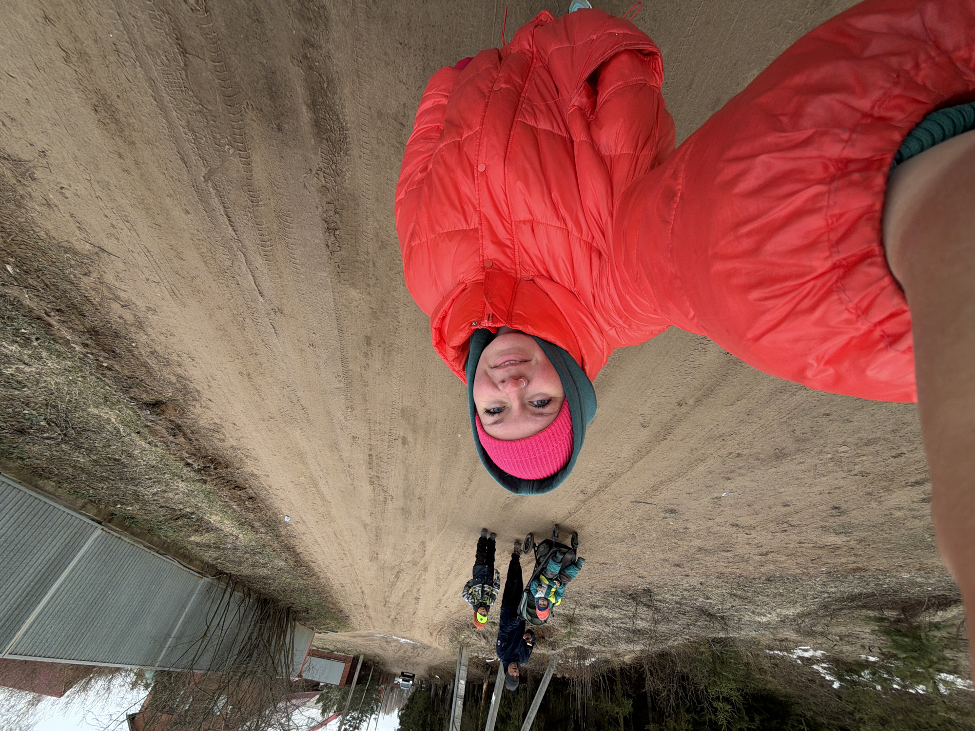 The image shows an individual wearing a red puffer jacket and pink beanie, appearing to take a selfie on a dirt road. Three additional individuals are visible further down the road, two standing and one in a stroller. A fence and some buildings are visible to the left, and trees to the right. Based on the description of being in Chaikovsky, Russia, this is likely a rural or semi-rural area.
