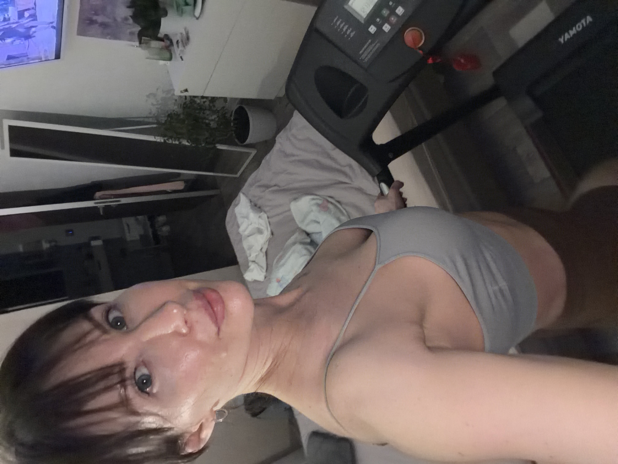 A woman is visible in a selfie taken indoors, likely in a domestic setting. She has light skin, dark hair, and is wearing a grey top. A treadmill is located behind her, and in the background, there is a television, a plant in a pot, and some furniture. The image provides no information to verify whether the photo was taken in Perm, Russia, as no indicators are present.