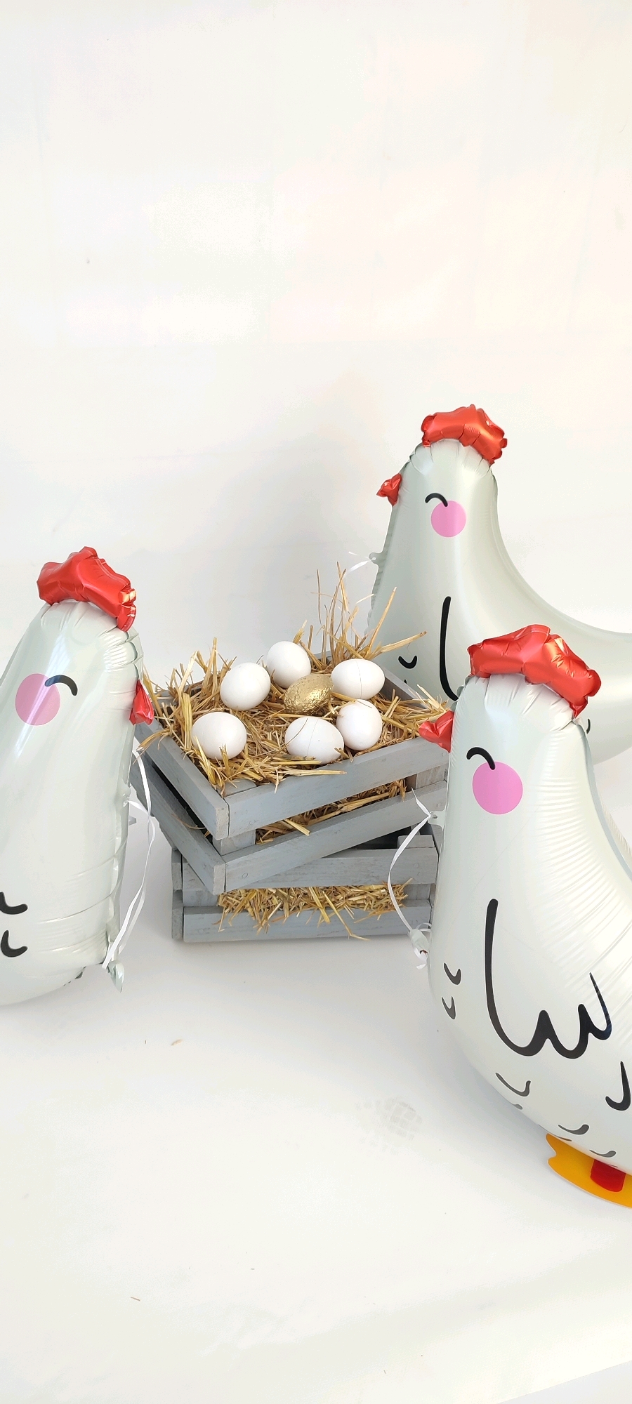 The image depicts a still life setup with four inflatable chickens and a wooden crate filled with straw and eggs. The chickens are white with red combs, and they have cartoonish faces drawn on them. The crate is gray and tilted, with several white eggs and one gold egg inside. The background is a plain white surface, suggesting a studio or indoor setting. There is no discernible connection to the city of Belaya Kalitva, Russia, as no identifying landmarks or contextual clues are present. The scene appears to be staged for a photograph or decorative display.