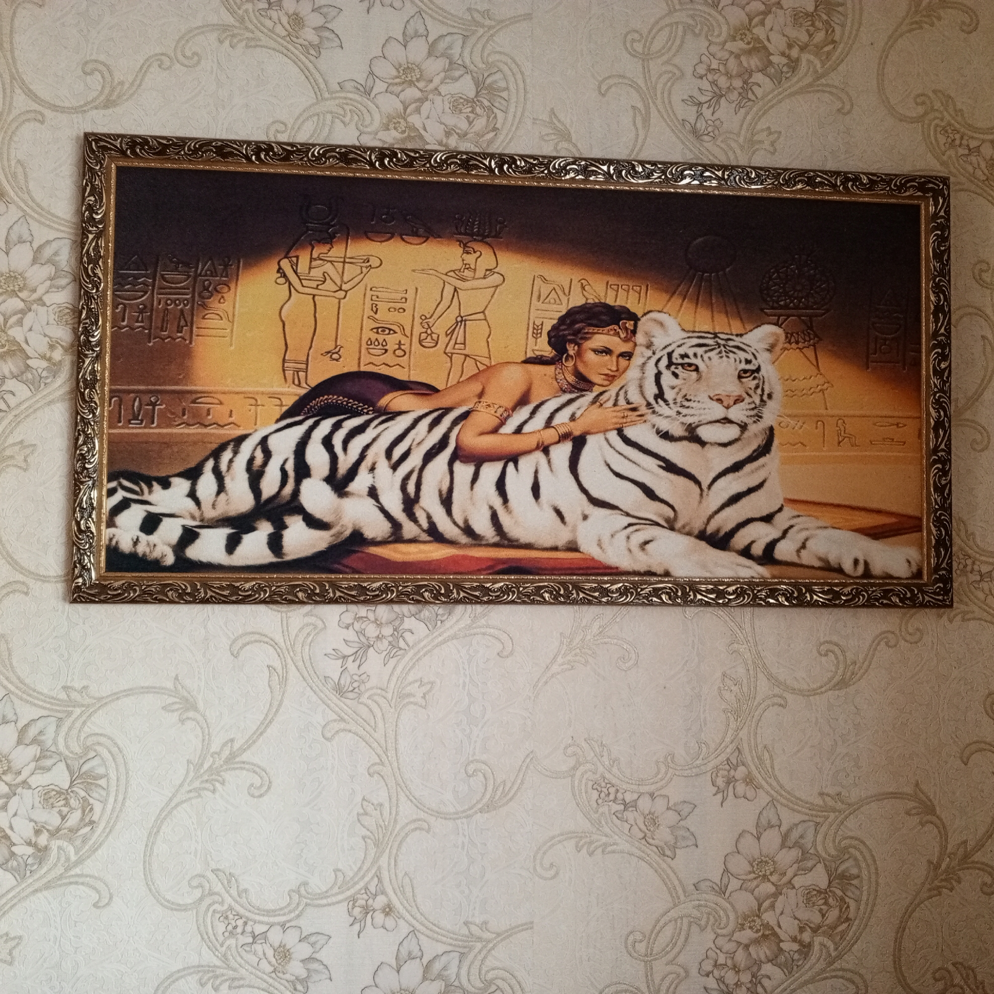 The image shows a framed painting hanging on a wallpapered wall. The painting depicts an ancient Egyptian scene with hieroglyphs and figures. In the foreground, a white tiger with black stripes is lying down. A woman with dark hair and Egyptian-style attire is reclining on the tiger's back, her arm resting on its neck. The wallpaper has a floral pattern. The painting is located on a wall in a room.