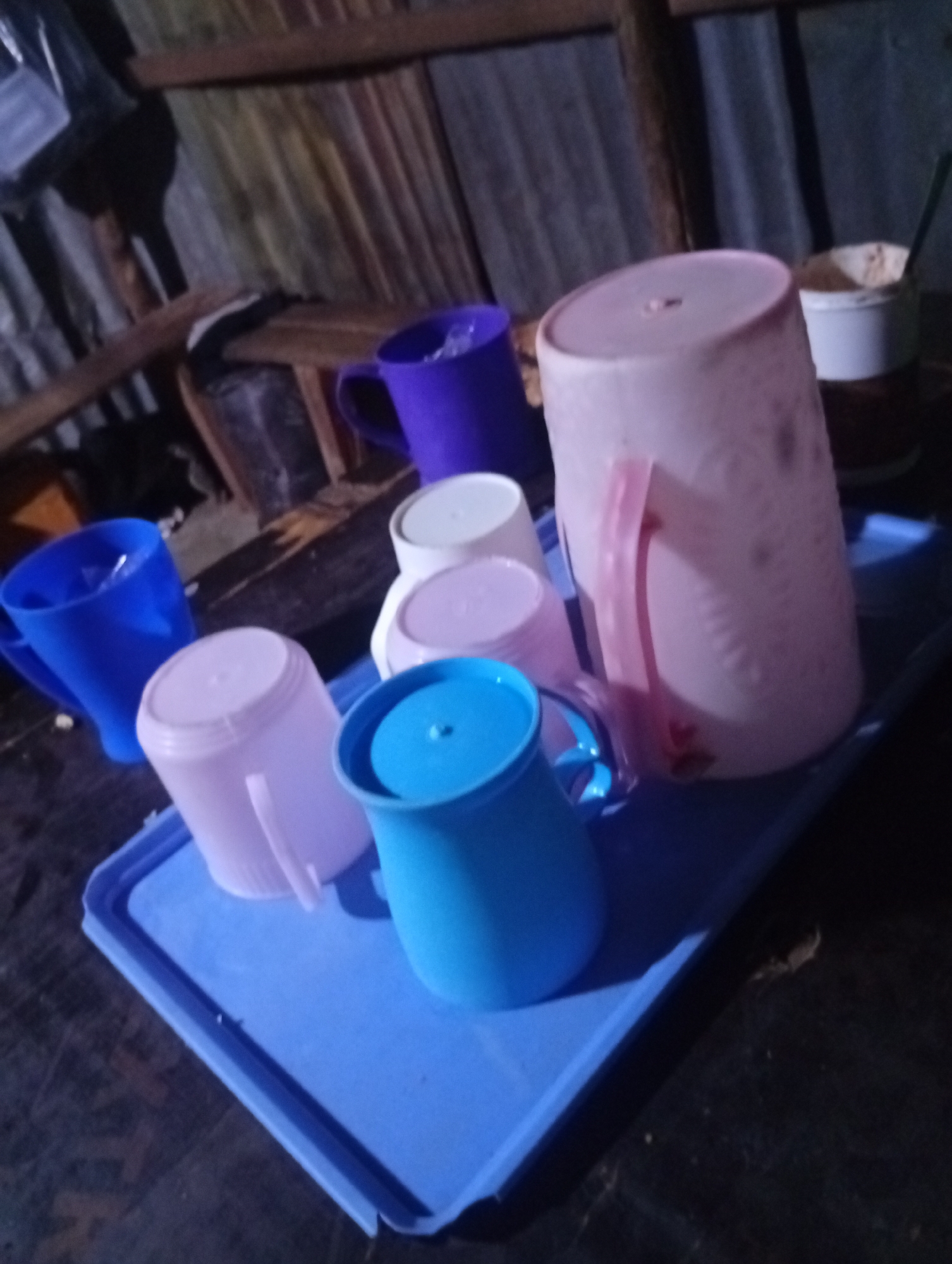 In an indoor setting with corrugated metal walls and wooden beams, a blue plastic tray holds six plastic cups of various colors. A large pink pitcher with a pink handle is the most prominent object. Next to it, a purple mug is visible. In front of these, three smaller cups are arranged: a pale pink, a white, and a light purple one. A bright blue cup with a lid is positioned in the foreground, also on the tray. A white cup containing a light-colored substance and a green utensil is seen on the far right, partially obscured. The lighting is dim, casting shadows and creating a somewhat grainy image.