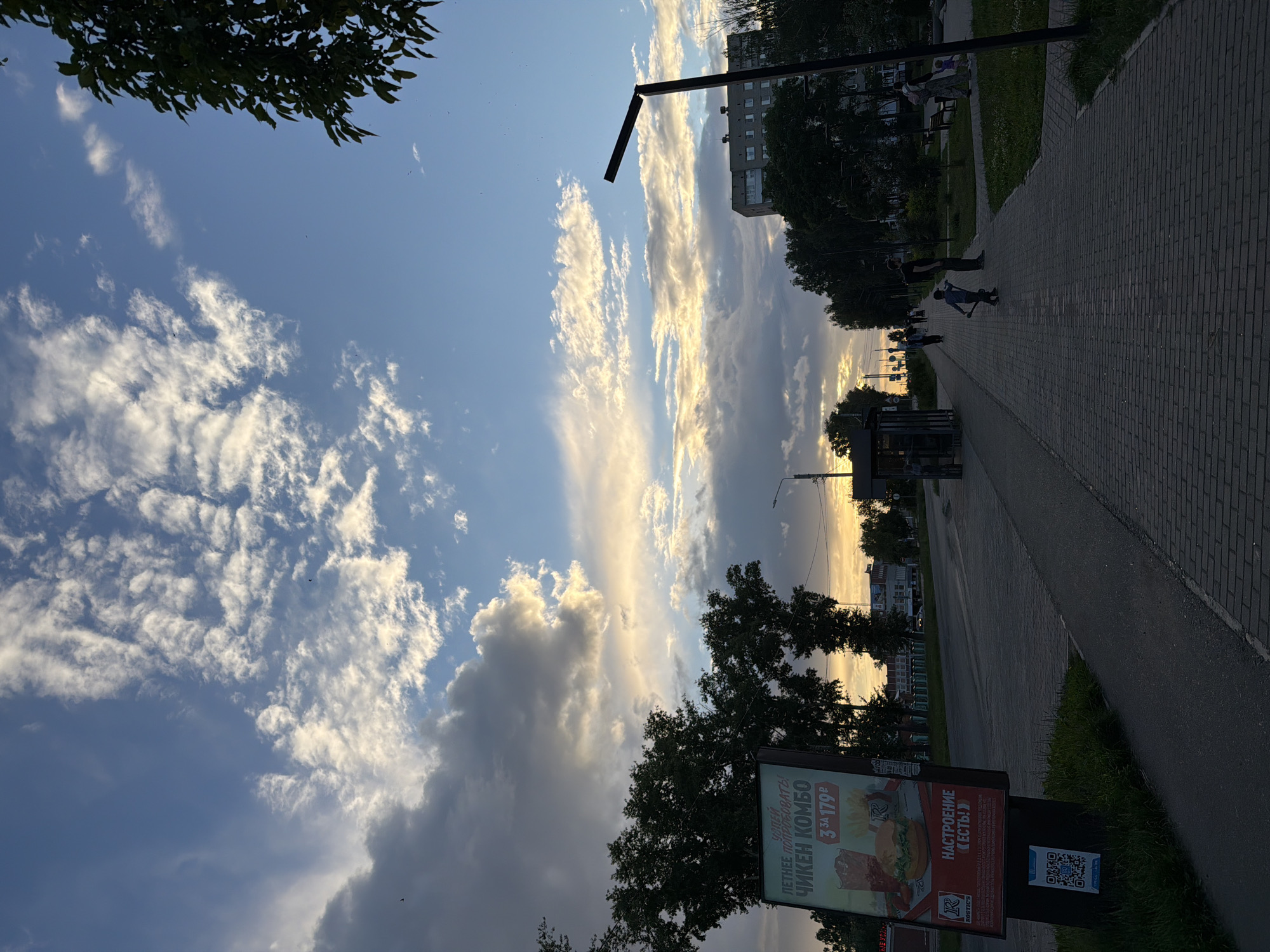This media file depicts a street scene in Chaikovsky, Russia, during what appears to be the late afternoon or early evening, given the warm, golden light of the setting sun illuminating the sky. The sky is a prominent feature, filled with dramatic clouds catching the sunlight, contrasting with patches of blue.
The scene is a paved walkway or sidewalk, bordered by a grassy area and trees. A tall, dark lamppost stands on the right side of the walkway. Further down the path, a few people are visible. One individual appears to be standing, while another is on a scooter, suggesting a leisurely activity. In the background, buildings and trees are silhouetted against the bright sky.
A large billboard advertising "ROSTIC'S" chicken combo is positioned on the left side of the image, near the bottom. The text on the billboard indicates a promotion: "ЛЕТНЕЕ ПОПРОБОВАТЬ! ЧИКЕН КОМБО 3 за 179 P" (Summer to try! Chicken combo 3 for 179 rubles) and "НАСТРОЕНИЕ «ЕСТЬ!»" (Mood "is there!"). A QR code is also visible on the billboard. The overall atmosphere seems peaceful, with a sense of everyday life unfolding as the day draws to a close.