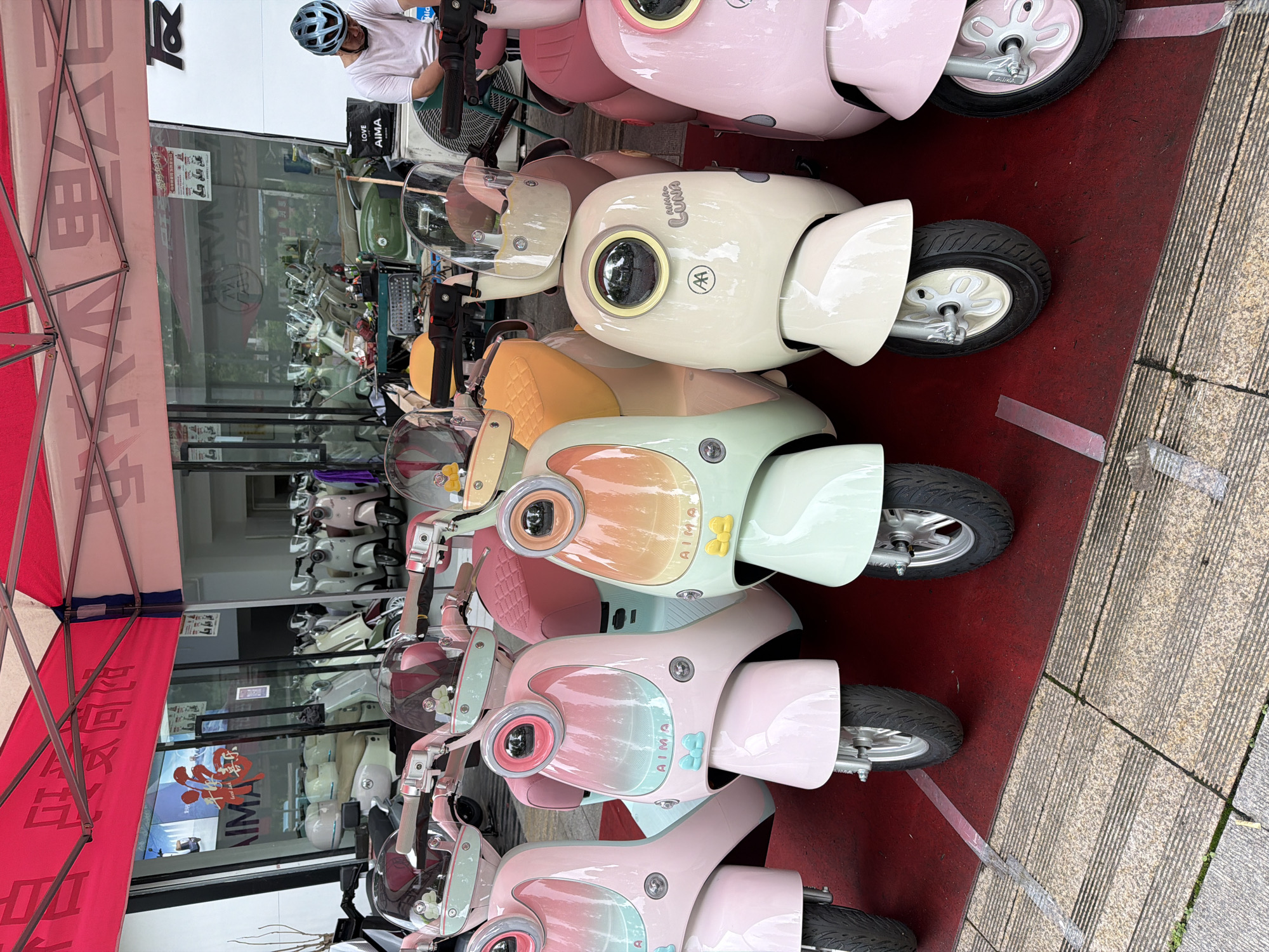 A row of Aima electric scooters, varying in pastel colors, are parked on a red mat next to a sidewalk composed of gray-striped tiles. The location is Chongqing High-tech Zone, China, as evidenced by the Chinese characters visible on an awning. A man wearing a helmet and a white shirt is sitting on the first scooter, seemingly observing or interacting with the display. Scooters are also reflected in the window behind this row of scooters.