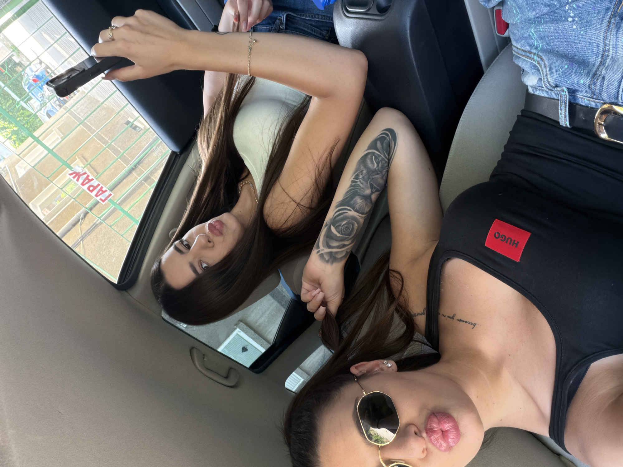 The image shows two young women inside a car. The woman in the foreground, on the left, is wearing a black tank top with a red "HUGO" logo, blue jeans, and a gold-buckled belt. She has long dark hair and is wearing sunglasses, with her lips pursed. She has tattoos on her arm and a small tattoo on her neck. The woman in the background, on the right, is wearing a white tank top and blue jeans. She has long dark hair and is holding a smartphone. The car's interior is visible, with grey seats and a dark dashboard. Through the car window, a building with tan-colored facade and green fencing is visible. A sign in Cyrillic script, reading "ГАРАЖ" (GARAGE), is attached to the fencing. This suggests the location is likely in Plovdiv, Bulgaria, given the Cyrillic signage. The overall lighting indicates it is daytime.