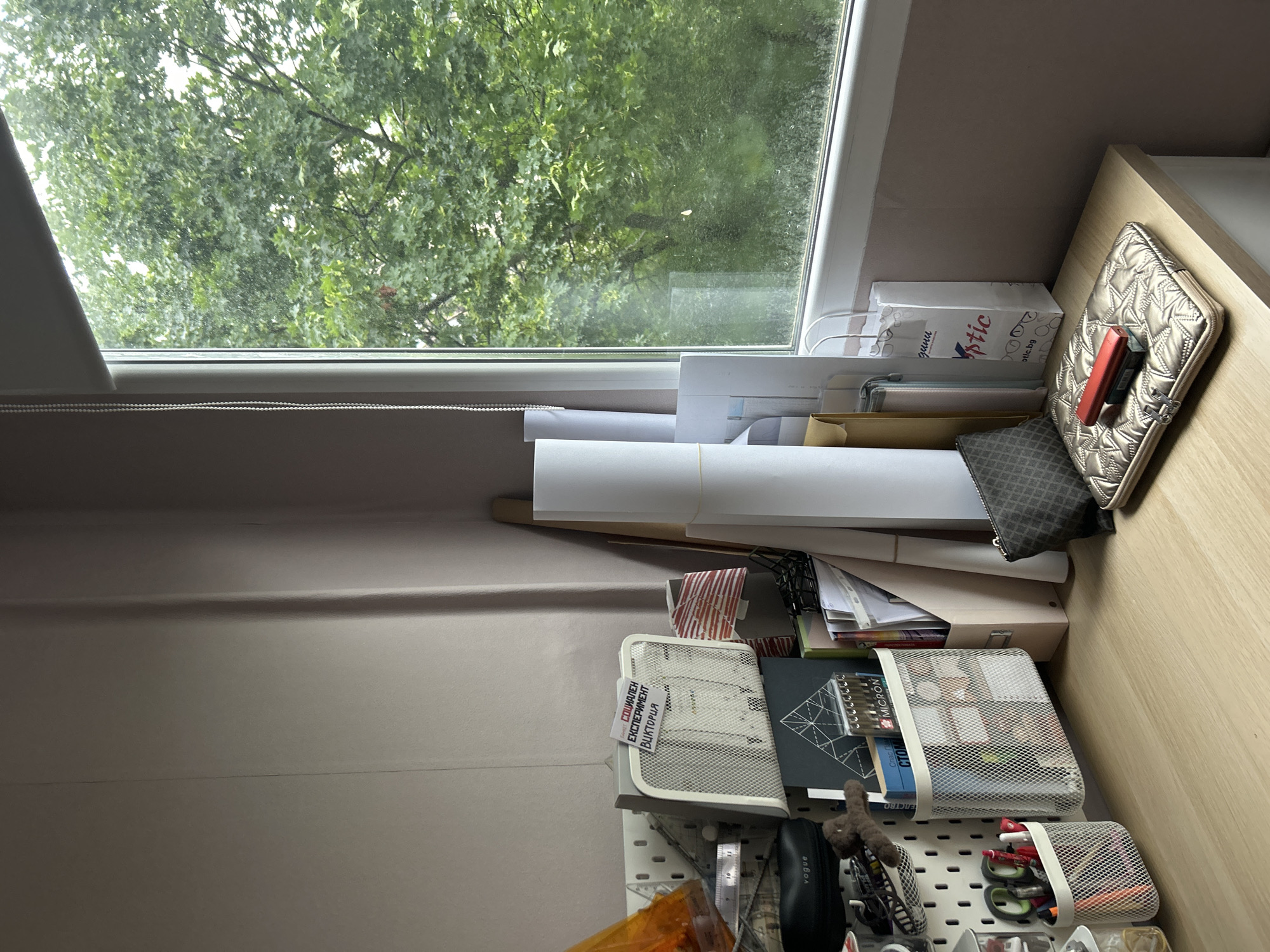This image captures a bright, cluttered workspace located indoors, with a window offering a view of lush green foliage outside. The scene is dominated by a desk or shelf area positioned beneath the window, organized with various items suggesting a creative or study environment.
Through the window, dense green leaves from trees are visible, indicating an outdoor natural setting, possibly a garden or park. The windowpane appears somewhat smudged or dusty. The natural light filtering through suggests it is daytime, possibly overcast, given the soft illumination.
On the right side of the image, a light wooden surface holds a quilted, metallic-colored laptop sleeve or case. A red, rectangular electronic device, possibly a portable hard drive or power bank, is tucked into the sleeve. Below the window, several large white rolled-up papers or canvases are stacked horizontally. Further to the left and below these, a white paper bag is visible, bearing the text "optic" and "optic.bg", along with a partial "gura". Other papers, folders, and small containers are also present.
A grey wall occupies the left side of the frame. Attached to it is a white pegboard system. Two white mesh containers hang on the pegboard, filled with assorted pens, markers, and other stationery items, including packaging with "MICRON" faintly visible. A black case, possibly for glasses, with the word "vogue" printed on it, is also attached. A small brown plush toy, a ruler, and a cutting mat displaying geometric patterns are discernible among the supplies. Above the pegboard, on the wall, there's a white rectangular object with a grid pattern, possibly a small fan or air purifier. A piece of paper taped to it clearly reads "СОЦИАЛЕН ЕКСПЕРИМЕНТ ВИКТОРИЯ", which translates from Bulgarian to "SOCIAL EXPERIMENT VICTORIA".
The presence of "optic.bg" and Bulgarian text aligns with the given location context of Sofia, Bulgaria. The arrangement of art supplies, papers, and electronic devices suggests an active creative or work-from-home setup.