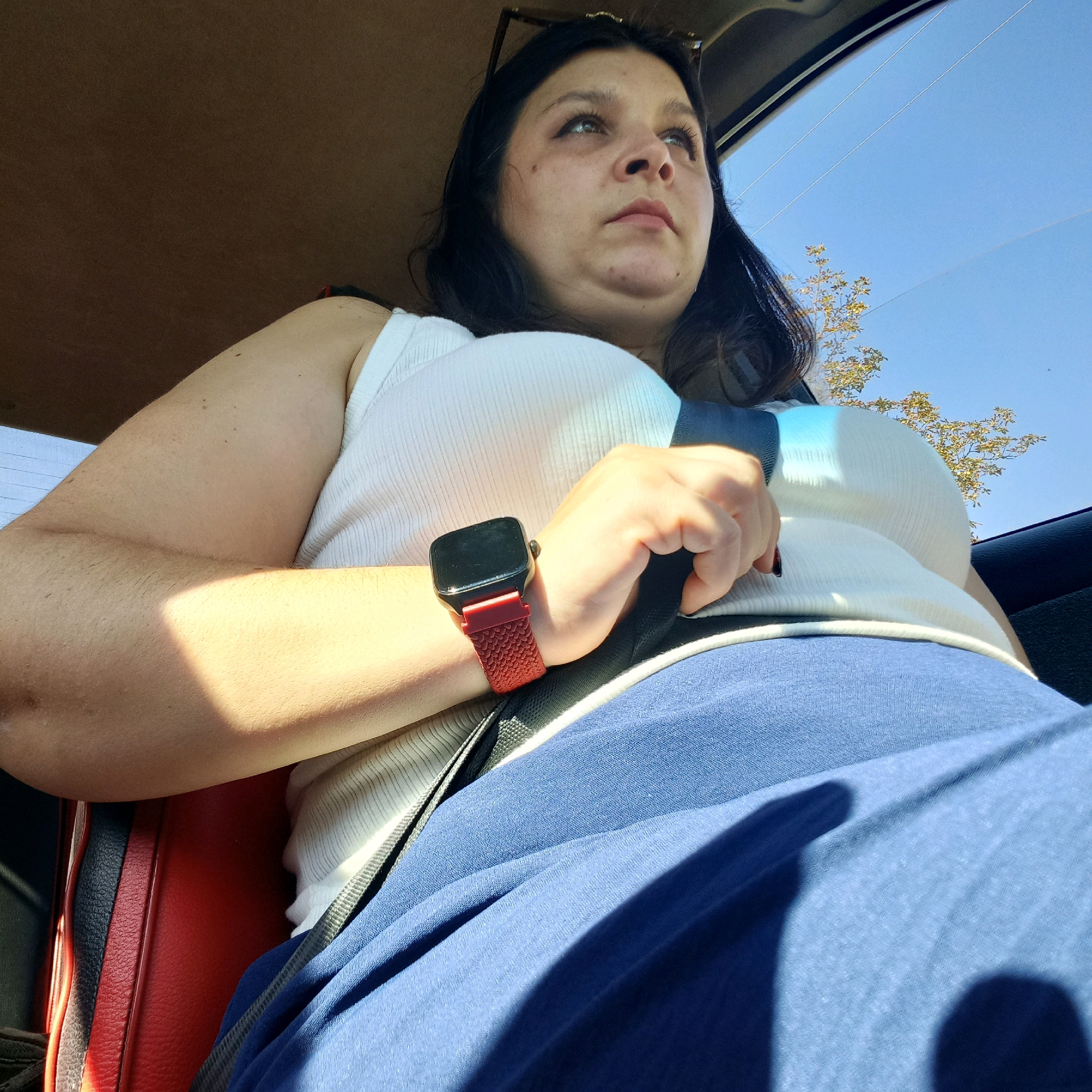 A woman with dark hair and fair skin is seated in the passenger seat of a vehicle, looking upwards and to the right. She is wearing a white ribbed tank top and a blue skirt. A seatbelt, partially obscured by her hand and arm, is visible across her torso. She is wearing a smartwatch with a red band on her left wrist. The interior of the car, including red upholstery, is visible. Sunlight streams in through the window, casting shadows. Outside the car, a clear blue sky and trees are visible. The image was taken in Klyntsi, Ukraine.