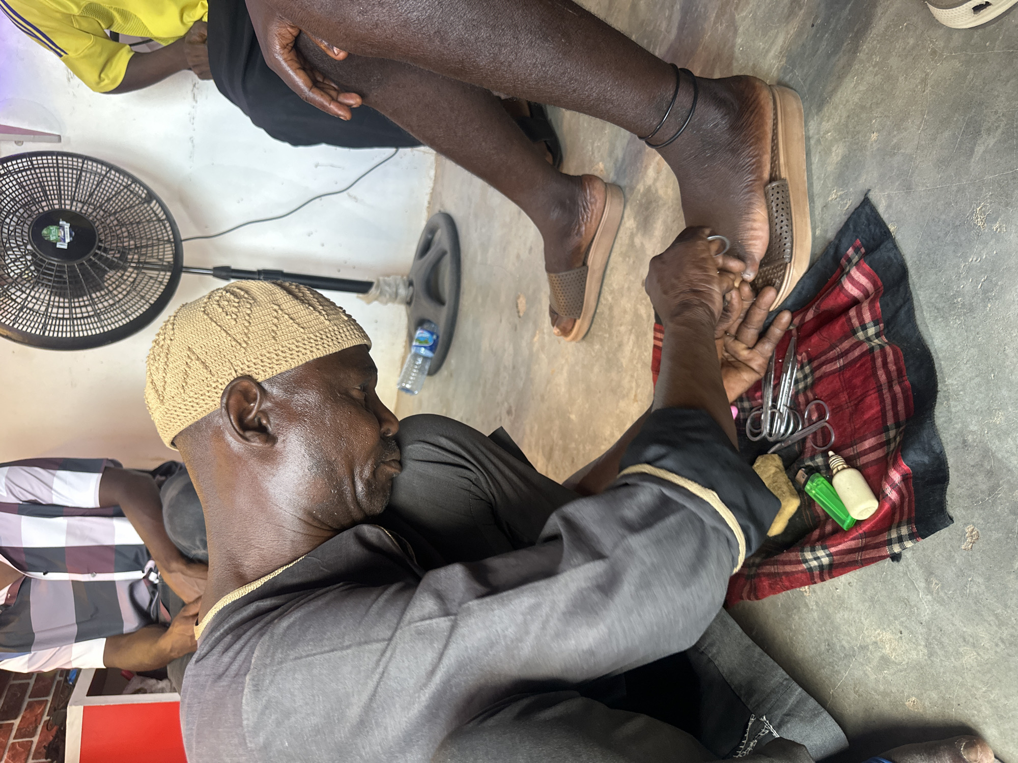 In a dimly lit room, a man in Jos, Nigeria, wearing a crocheted cap, sits on the floor, attending to the toenails of a seated person. The person being attended to wears an ankle bracelet and brown sandals. A checkered cloth on the floor holds grooming tools: scissors, lighters, and what appear to be bottles of solution. An electric fan stands nearby.