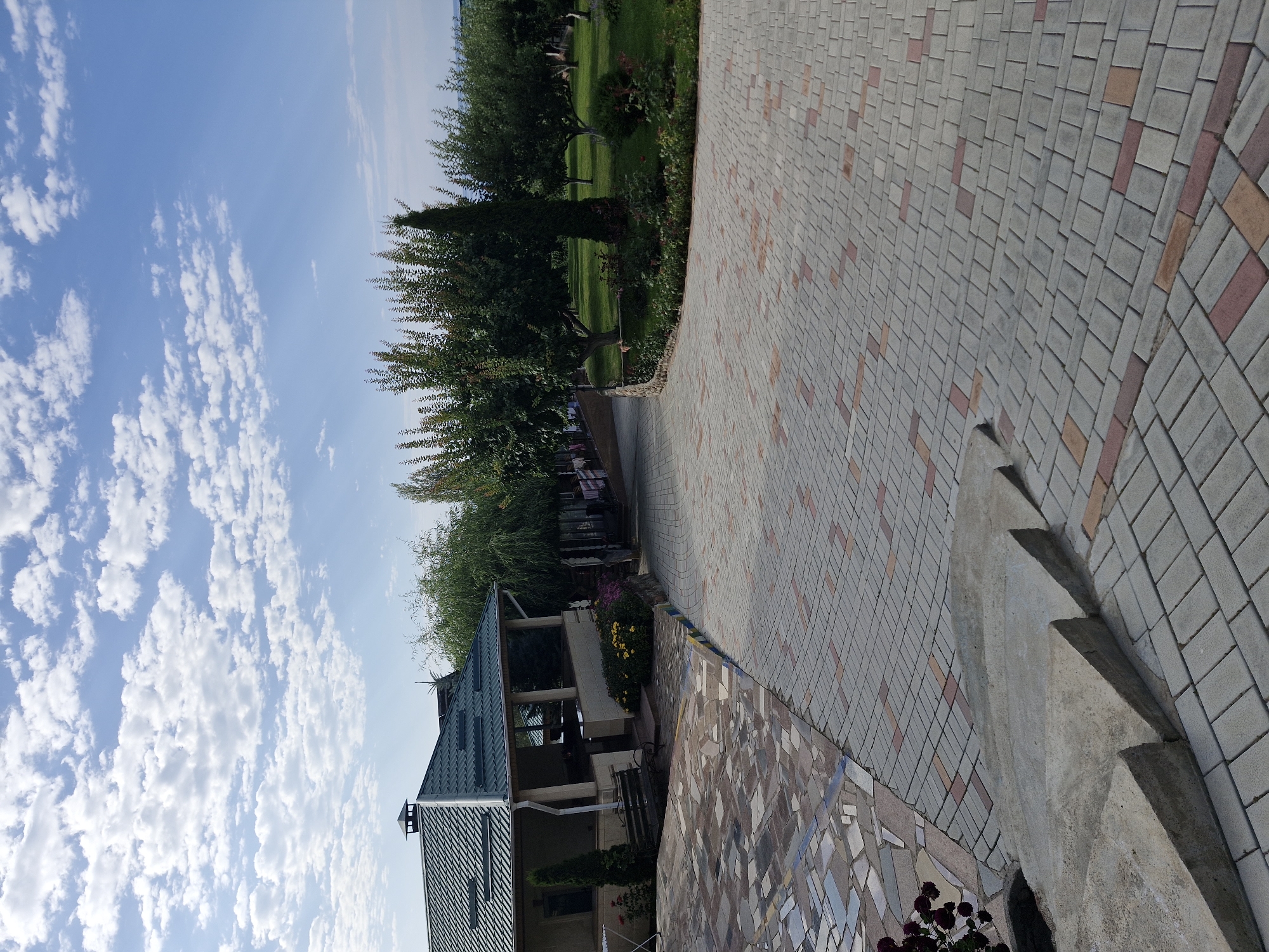 The image shows an outdoor paved area with a building and a garden in the background. The paving is made of rectangular bricks in shades of grey, red, and pink, arranged in a pattern. In the foreground, there are concrete steps. To the left, a section of the paving is made of irregular-shaped stones in various colors. A building with a green metal roof and an open-air veranda is visible on the left. In front of the building, there is a bench and some plants. The background features a well-maintained garden with trees, grass, and flowers. The sky above is blue with scattered white clouds. The scene appears to be a resort or a private residence in Bulan-Segettu, Kyrgyzstan. There are no people visible in the image.