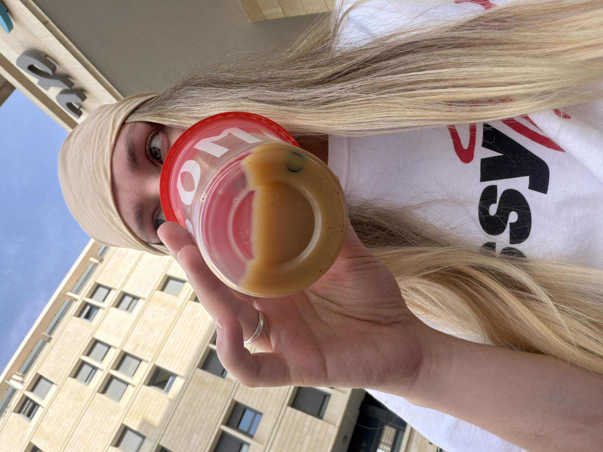 A fair-skinned woman with long, blond hair and a beige headband is holding a clear plastic cup with a red lid containing a liquid. The cup is partially filled with two different-colored liquids, one red and one yellow-brown. The woman is wearing a white t-shirt with the word "MISS" printed on it in black and red lettering. Her left hand is visible, with a silver ring on her ring finger. The background features a light yellow building with many windows and a blue sky. The photograph was taken in Tel Aviv, Israel.