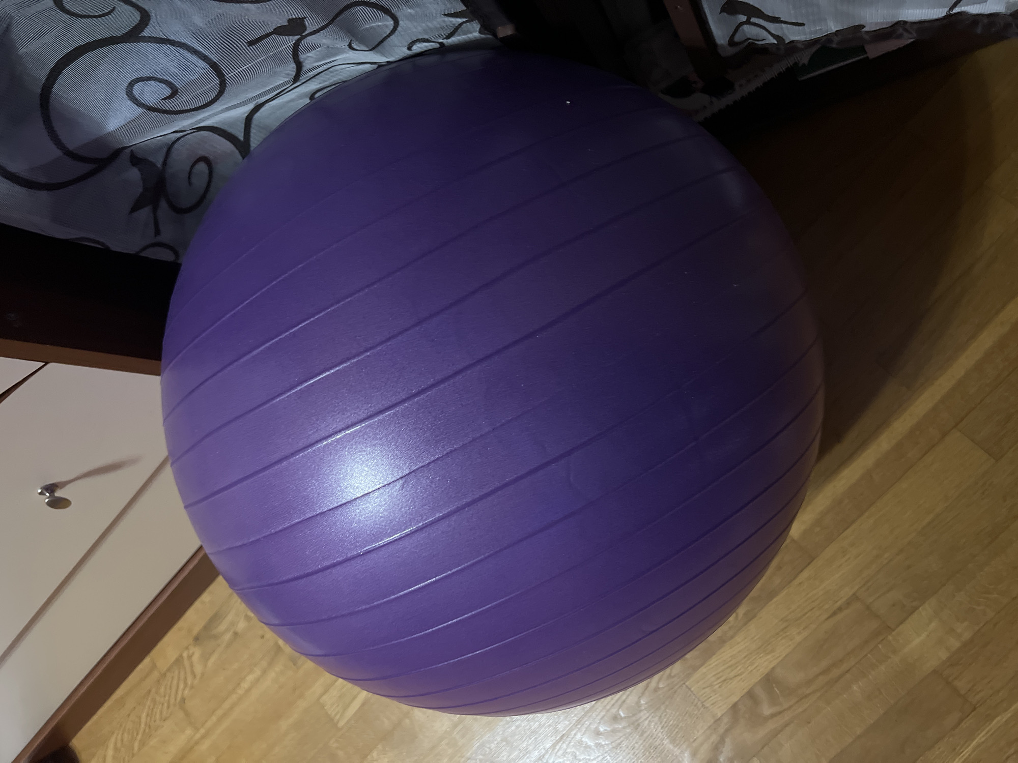 A large, purple exercise ball is positioned on a wooden floor. The ball is textured with parallel grooves. To the left of the ball, part of a white drawer unit with a silver knob is visible. To the right, a dark wooden structure and a patterned fabric are present. The scene appears to be indoors, possibly a room in an apartment or house. There is no indication of the specific location within Minsk, Belarus.