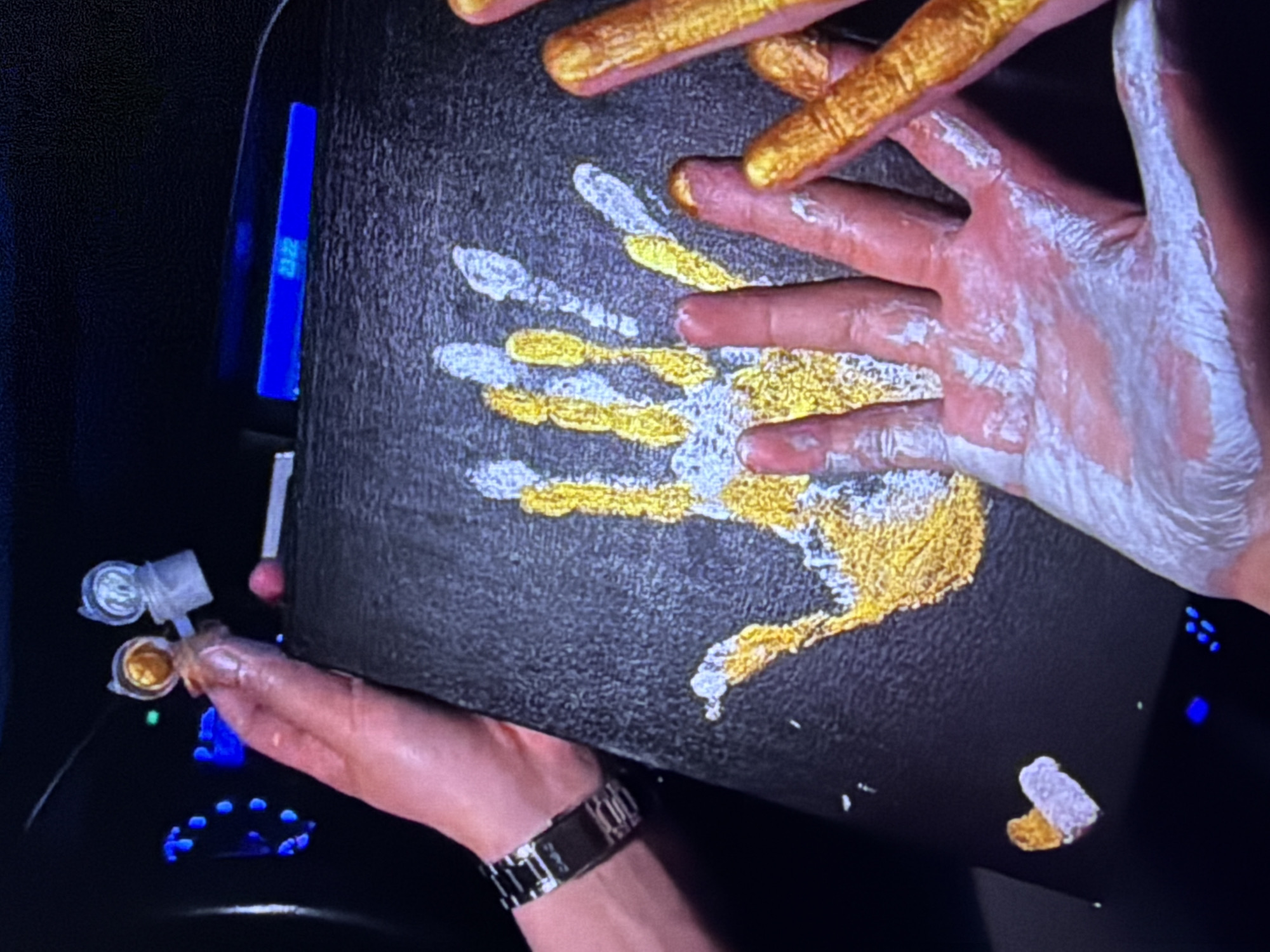 In Beșalma, Moldova, an individual is creating a handprint artwork on a dark canvas. The canvas has a large handprint impressed on it, with fingers outlined in gold and white paint. A second hand, also painted gold, is positioned to the right of the canvas. The person's left hand, coated in white paint, is in the foreground. In the background, a digital display shows "23:22," possibly indicating the time. Two small bottles, one containing gold paint and the other an unknown substance, are visible near the top left. A small heart shape, half gold and half white, is present on the canvas near the bottom left. The scene appears to be indoors, with artificial lighting contributing to the ambiance.