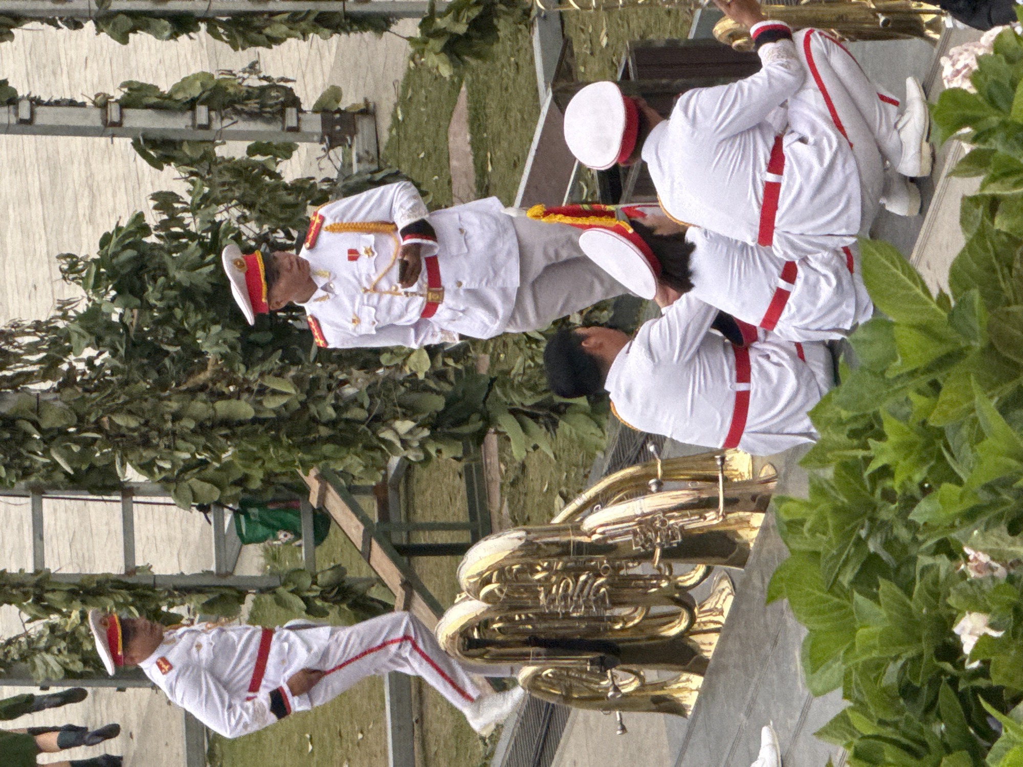 This media file captures a group of people in uniform, seemingly participating in a parade or performance. The subjects are dressed in white uniforms with red trim, including hats, jackets, and pants. Several individuals are holding brass instruments, specifically tubas, suggesting a musical ensemble. The scene appears to be outdoors, with foliage and what looks like a structure in the background, possibly a stage or seating area. The lighting suggests it could be daytime. The overall atmosphere seems formal and organized, with the participants maintaining a composed posture. The backdrop includes greenery and a metallic structure. There is no visible text in the image. The context suggests this event is taking place in Da Lat, Vietnam, a location known for its pleasant climate and outdoor gatherings.