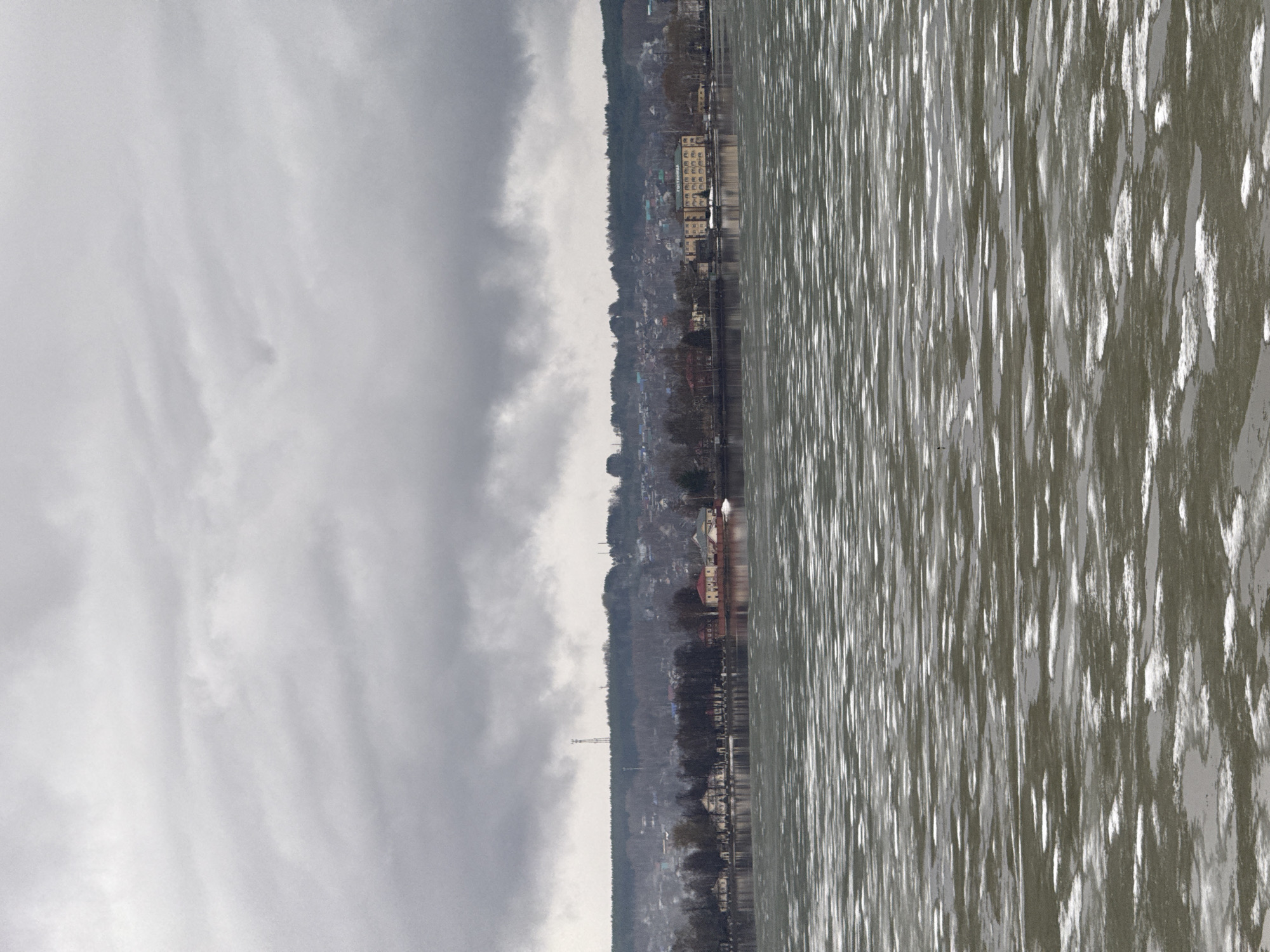 The image captures a water body, likely a reservoir or river, adjacent to a shoreline dotted with buildings and a line of trees, possibly within or near the city of Votkinsk, Russia. The sky is overcast, with a dense layer of grey clouds obscuring the horizon. The water surface reflects the ambient light, creating a shimmering effect with streaks of light and dark tones. A mix of residential and commercial buildings is visible along the shoreline, interspersed with vegetation. A tall communications tower is partially visible. The overall impression is of a calm, overcast day with the interplay of natural and built environments near a body of water.