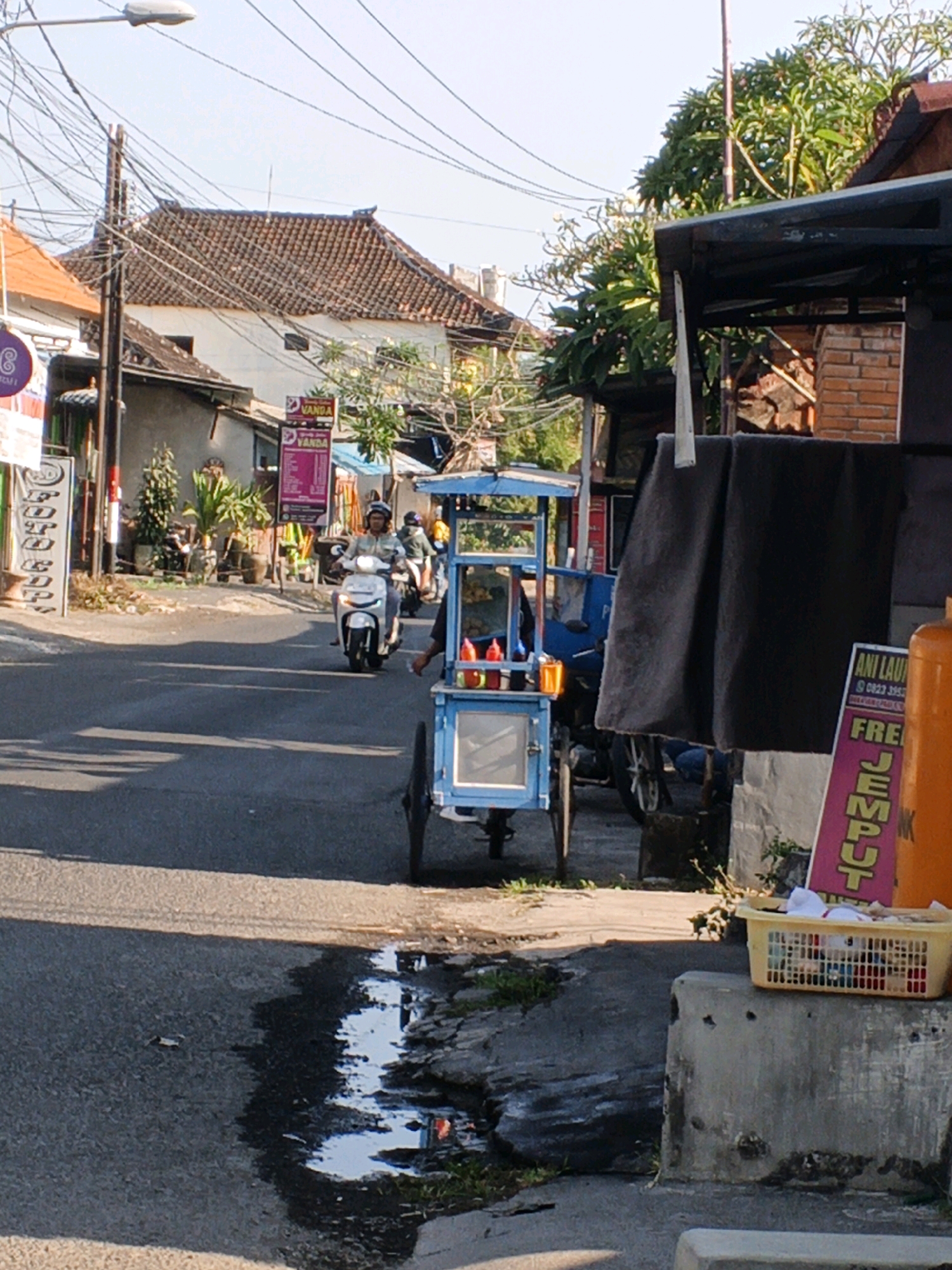 The image depicts a street scene in Sukawati, Indonesia, on a sunny day. A person wearing a helmet rides a white scooter, heading towards the viewer. To the right, a blue food cart, likely for selling snacks or drinks, is parked. The cart has a clear display window and two red sauce bottles on top. Behind the cart, a dark gray fabric hangs from a structure. Further to the right, a pink sign with yellow text is visible, advertising something with the word "JEMPUT." A yellow basket containing white items is placed on a concrete block in the foreground. The street is paved, and a small puddle of water reflects the sky. In the background, buildings with tiled roofs, trees, and utility poles with numerous wires are present. The overall atmosphere suggests a typical day in a bustling Indonesian town.