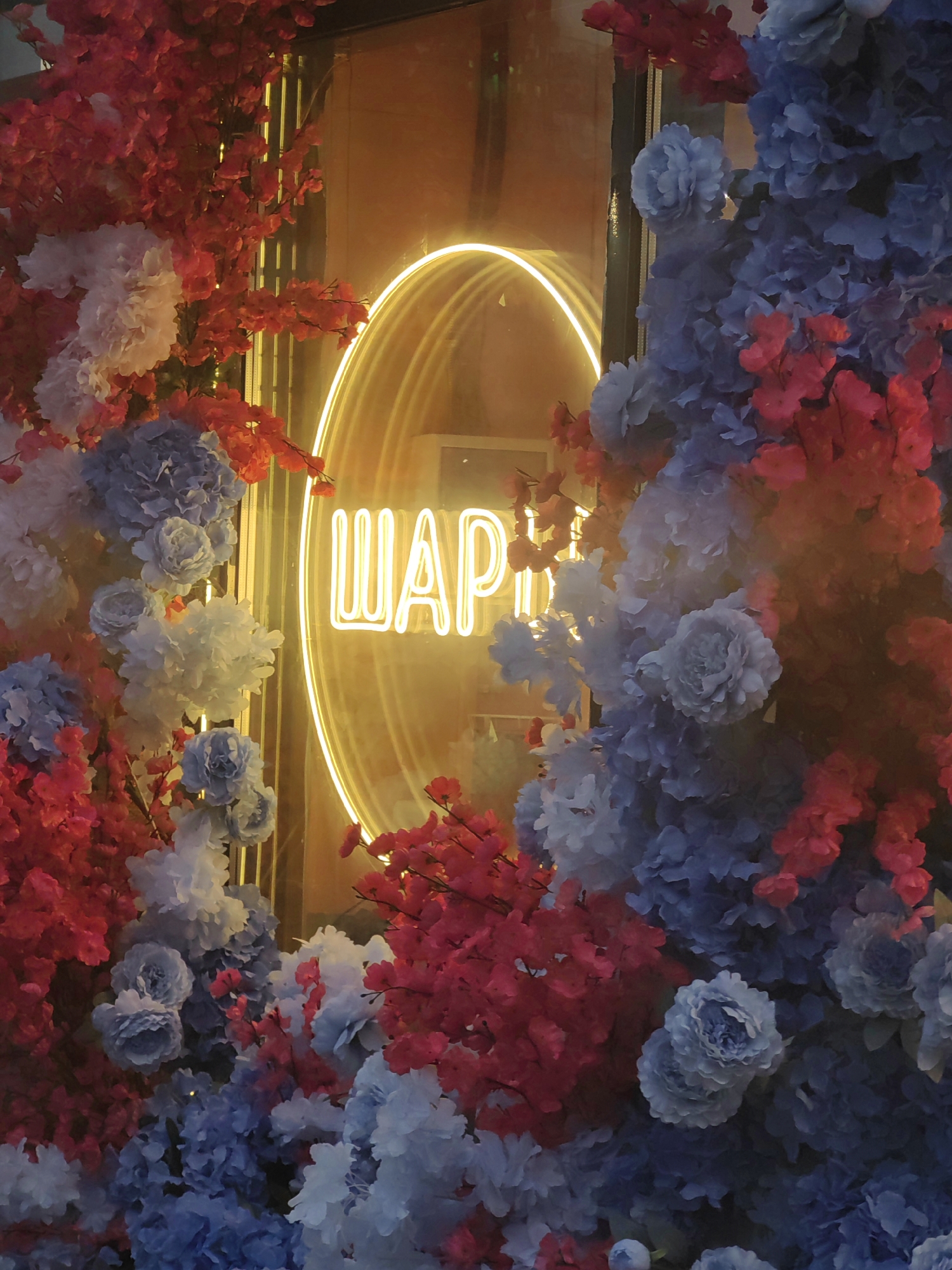 The image depicts a window display, likely in the city of Mytishchi, Russia. A bright yellow neon sign, forming a circle and displaying the word "ШАР" (translated to "SHAR" or "BALL" in English), is centrally located. The sign is surrounded by artificial floral arrangements, featuring predominantly red and blue blossoms. These flowers frame the neon sign, adding a decorative element to the display. The lighting suggests either dusk or nighttime. The scene is focused on the shop front and its decorative elements.