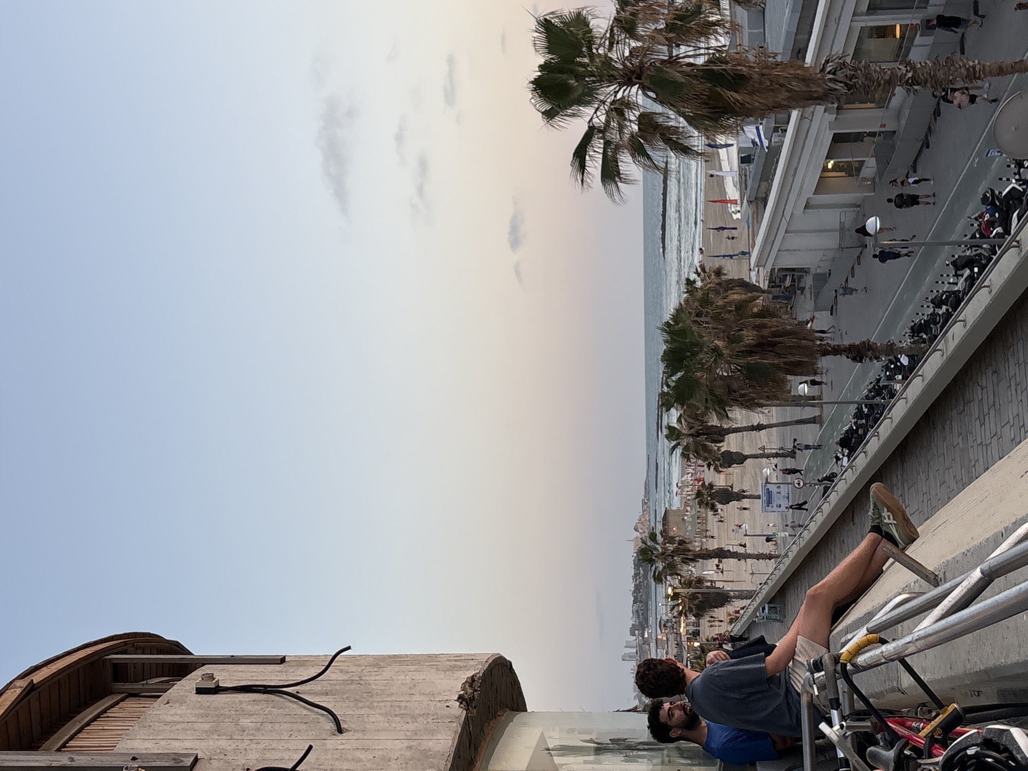 The image captures a relaxed coastal scene in Tel-Aviv, Israel, during late afternoon or early evening, viewed from an elevated perspective.
In the foreground, two men are visible on what appears to be a balcony or elevated platform. One man sits casually with his legs propped up on a metal railing or scaffolding, wearing shorts, a dark t-shirt, and green-soled sneakers. Beside him, another man in a blue t-shirt leans forward, looking out over the scene. Parts of bicycles are also visible near them.
Below and stretching horizontally across the middle of the frame is a wide promenade, busy with pedestrians strolling along. It is lined with numerous tall palm trees and modern buildings. A row of parked motorcycles or scooters can be seen along the side of the promenade.
To the left of the promenade lies a sandy beach, dotted with several colorful beach flags or umbrellas. The Mediterranean Sea extends beyond the beach, with gentle waves breaking on the shore. In the far distance, a faint city skyline is visible along the coastline.
The sky, transitioning from a pale blue directly overhead to a warmer, soft yellow-orange closer to the horizon, indicates the time of day is dusk or twilight. A few wispy clouds are scattered across the sky, contributing to the tranquil atmosphere. The weather appears clear and calm. No legible text is discernible on any visible signs.