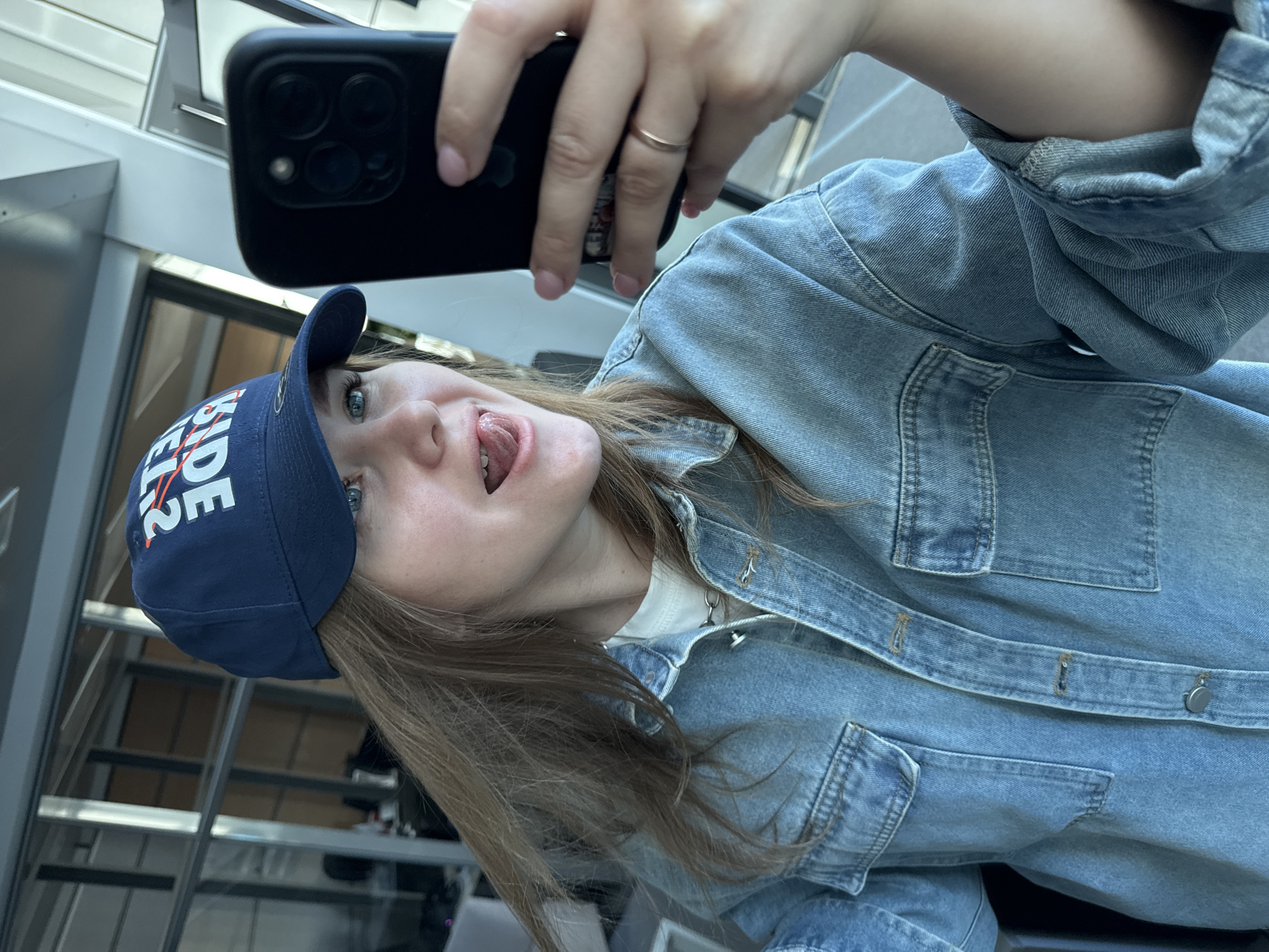 A person is holding a black iPhone in their right hand, taking a selfie from a low angle. The person is wearing a blue baseball cap with white lettering, a light blue denim jacket, and a white shirt underneath. They are sticking their tongue out. A silver ring is on the ring finger of their right hand. The background is a building with metal framing and glass panels. The location is in Perm, Russia.