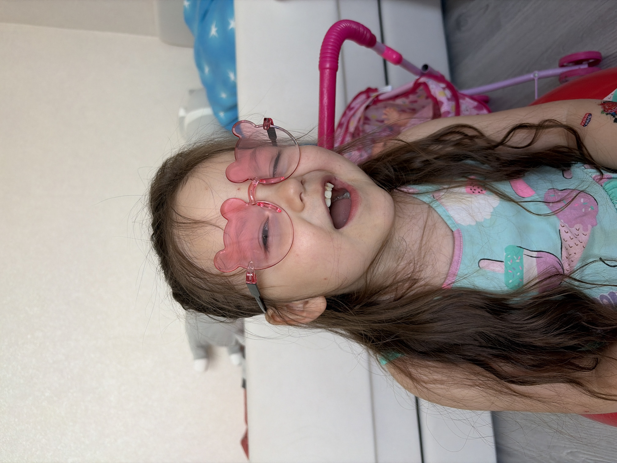 A young girl with light skin and brown hair is lying on her side, wearing pink bear-shaped sunglasses and a light blue patterned tank top. Her mouth is open in a smile. A pink toy stroller is near her head. The location cannot be determined as Izhevsk, Russia. The background includes a white wall and a gray floor. A stuffed animal appears in the bottom left corner.