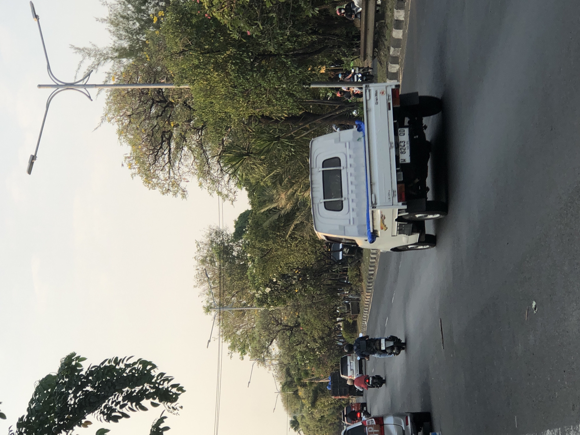 In Ngaban, Indonesia, a white pickup truck with license plate W 6243 QD is driving on a paved road. The truck is in the foreground, facing away from the camera. The road is lined with trees and streetlights. There are other vehicles on the road, including motorcycles and cars. Some people are riding motorcycles, and one person is sitting in the passenger seat of a car. The sky is overcast, and the sun is setting.