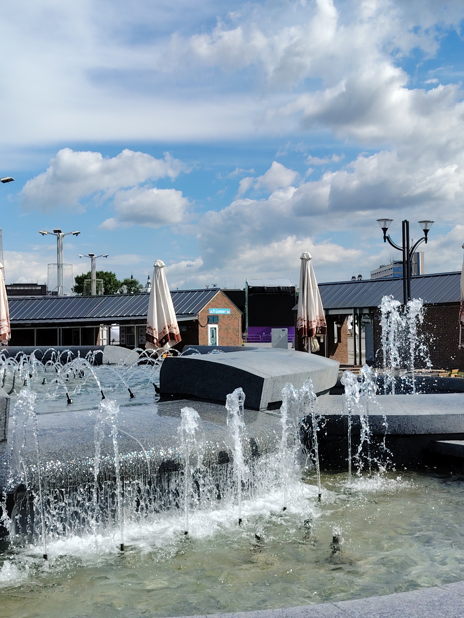 The media file captures a bright, sunny daytime scene featuring an active modern fountain in an urban setting, likely a public plaza in Minsk, Belarus.
In the foreground, a multi-tiered fountain made of dark gray stone or concrete features numerous jets spraying water upwards and cascading over ledges into pools below. The water appears clear, with sunlight reflecting off the droplets and the surface.
Behind the fountain, a row of low-rise buildings with dark, sloped metal roofs and some brick facades are visible. Several large, cream-colored umbrellas (parasols) are folded and positioned near these buildings, suggesting outdoor cafe seating areas that are currently not in use. One umbrella prominently displays a faint 'K' in a dark pattern. On a brick building to the right, a small blue sign with illegible white text and a white sign below it are visible.
The background is dominated by a bright blue sky dotted with fluffy white cumulus clouds. Modern-style lampposts with multiple light fixtures rise above the fountain and buildings. Some green foliage and trees are seen peeking over the roofs in the distance, along with glimpses of other taller city structures.
The overall impression is one of a peaceful, well-maintained public space on a clear, pleasant day. No people are visible in the image, emphasizing the architectural and natural elements.