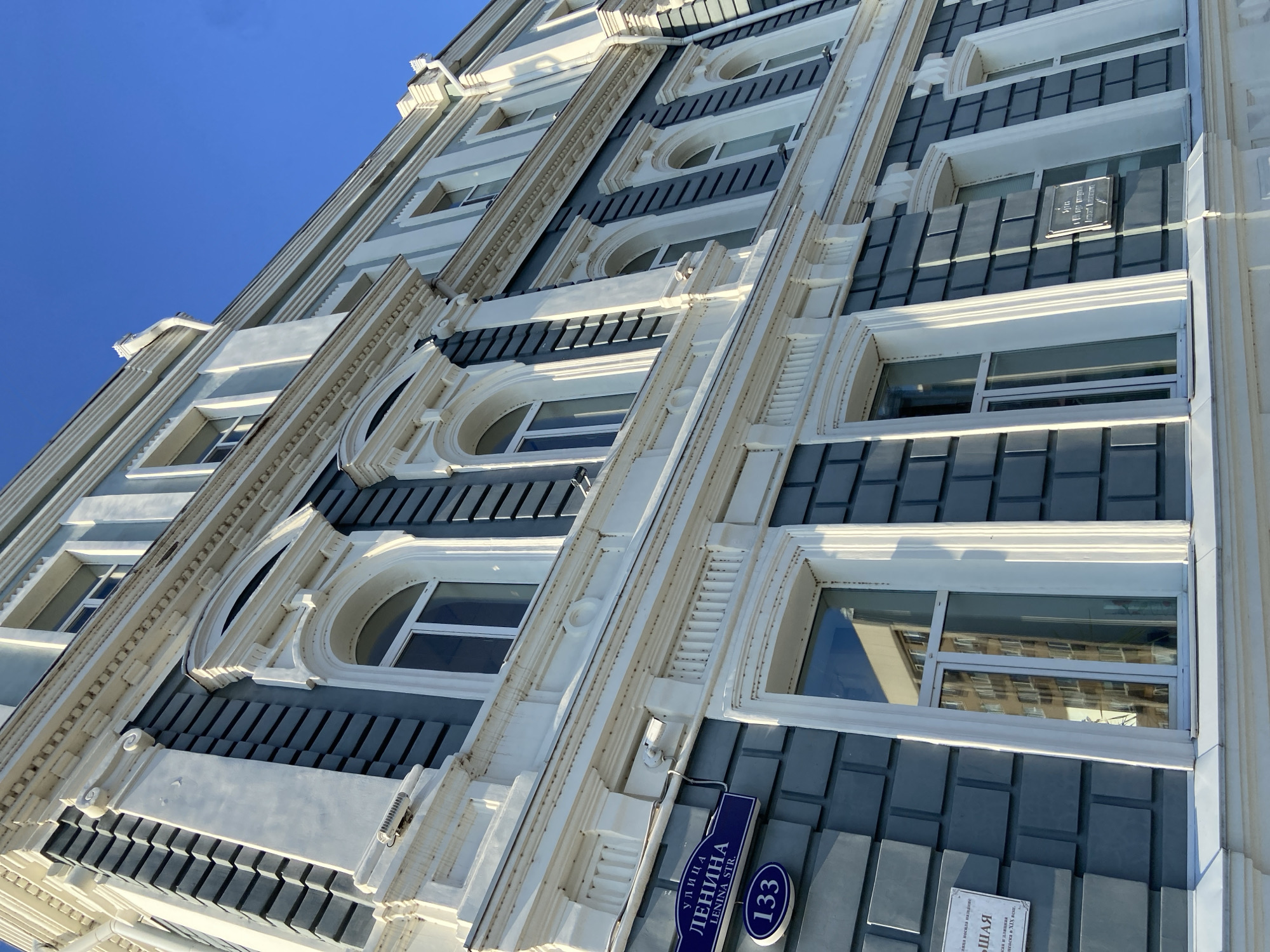 The image shows the exterior of a building with a light blue facade and white decorative elements, likely in the city of Blagoveshchensk, Russia. The building features multiple windows, some arched and some rectangular, with white frames. A blue street sign that reads "УЛИЦА ЛЕНИНА" (Ulitsa Lenina) and "LENINA STR." with the number "133" is visible on the left side of the building. Another plaque with text in Russian is also present. The building is viewed from a low angle, looking upwards against a clear blue sky.