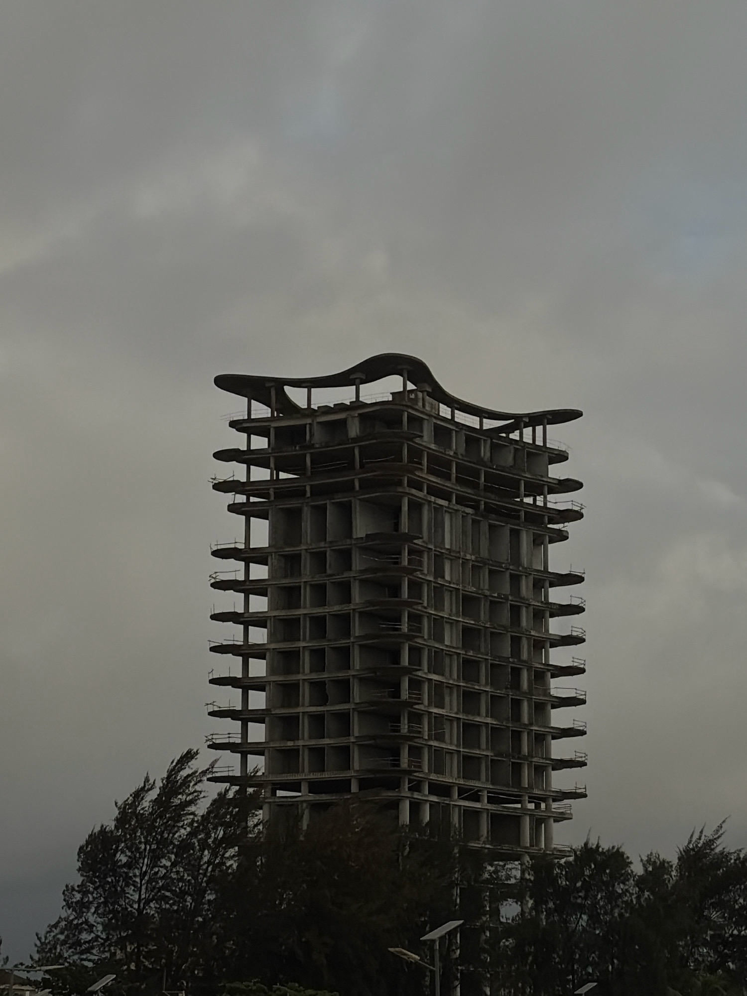 An unfinished, multi-story concrete building stands against a cloudy sky in Alaguntan, Nigeria. The building's structure is exposed, revealing floors with open spaces and visible beams and columns. Balconies or external walkways are present on several levels, some with rudimentary safety railings. The top of the building features a distinct, curved roofline. In the foreground, the tops of dark green trees are visible, some appearing to be wind-swept. The overall scene is somber, likely due to the overcast weather and the state of the construction.