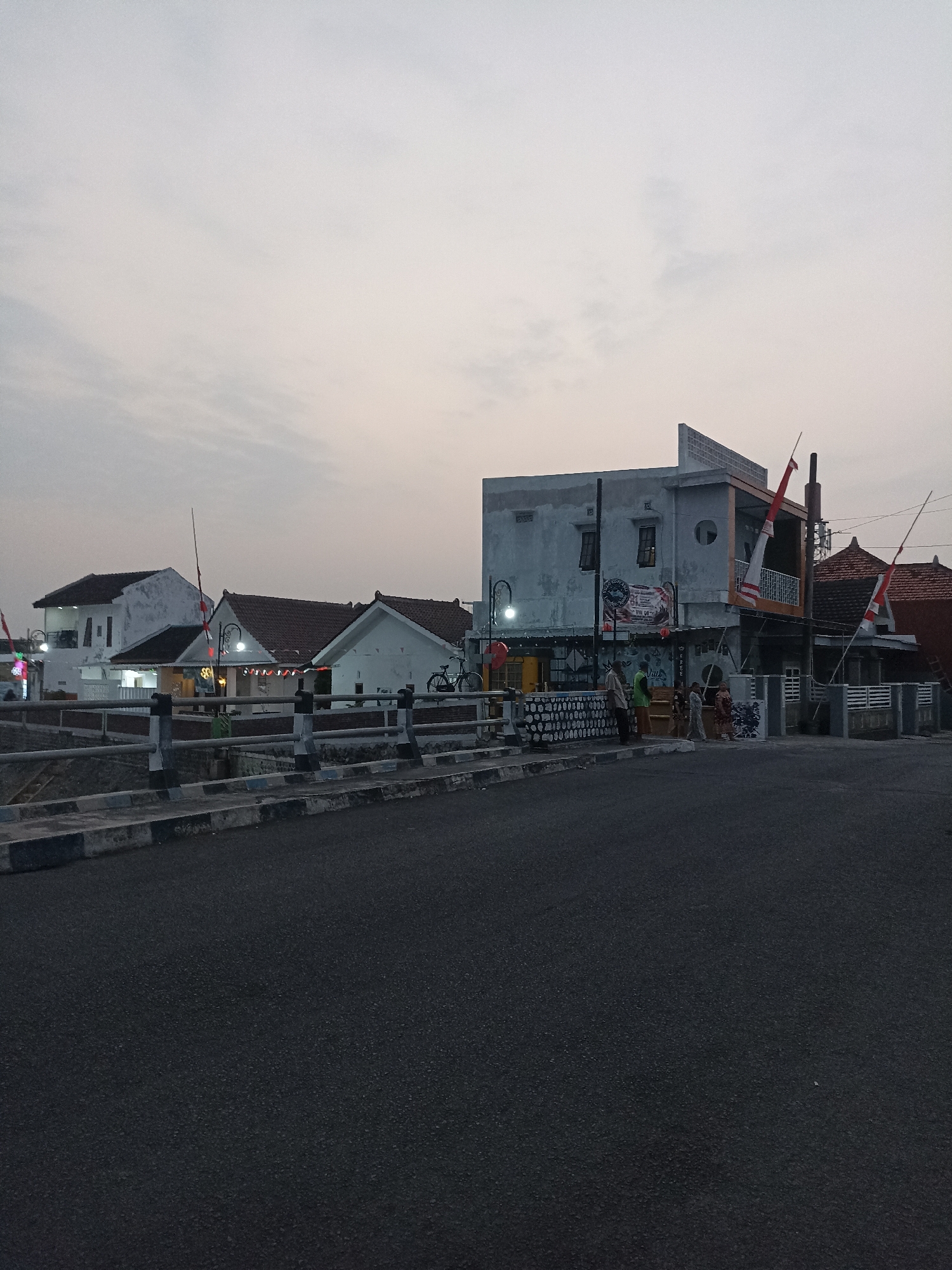 This image captures a street scene in Lumajang, Indonesia, during dusk. In the foreground, a paved road runs from the bottom of the frame upwards and slightly to the left. Along the right side of the road, a concrete curb with black and white markings is visible. A low concrete barrier with black and white bollards separates the road from a canal or drainage ditch.
Beyond the barrier, several buildings are visible. On the left, a two-story white building with a dark roof has windows and lights illuminating its interior. To its right, a lower, single-story white building with a reddish-brown tiled roof features several windows and decorative elements, along with several Indonesian flags displayed prominently on poles. Further to the right, a more modern, multi-story building with a gray facade and an orange accent on the upper story stands out. This building also displays Indonesian flags. Several people are standing near this building, some appearing to be customers or employees. A bicycle is parked near the single-story building on the right.
The sky above is overcast with soft, diffused light, indicating either sunrise or sunset. The overall atmosphere of the scene is quiet and typical of a residential or commercial street in a provincial town at the end of the day.