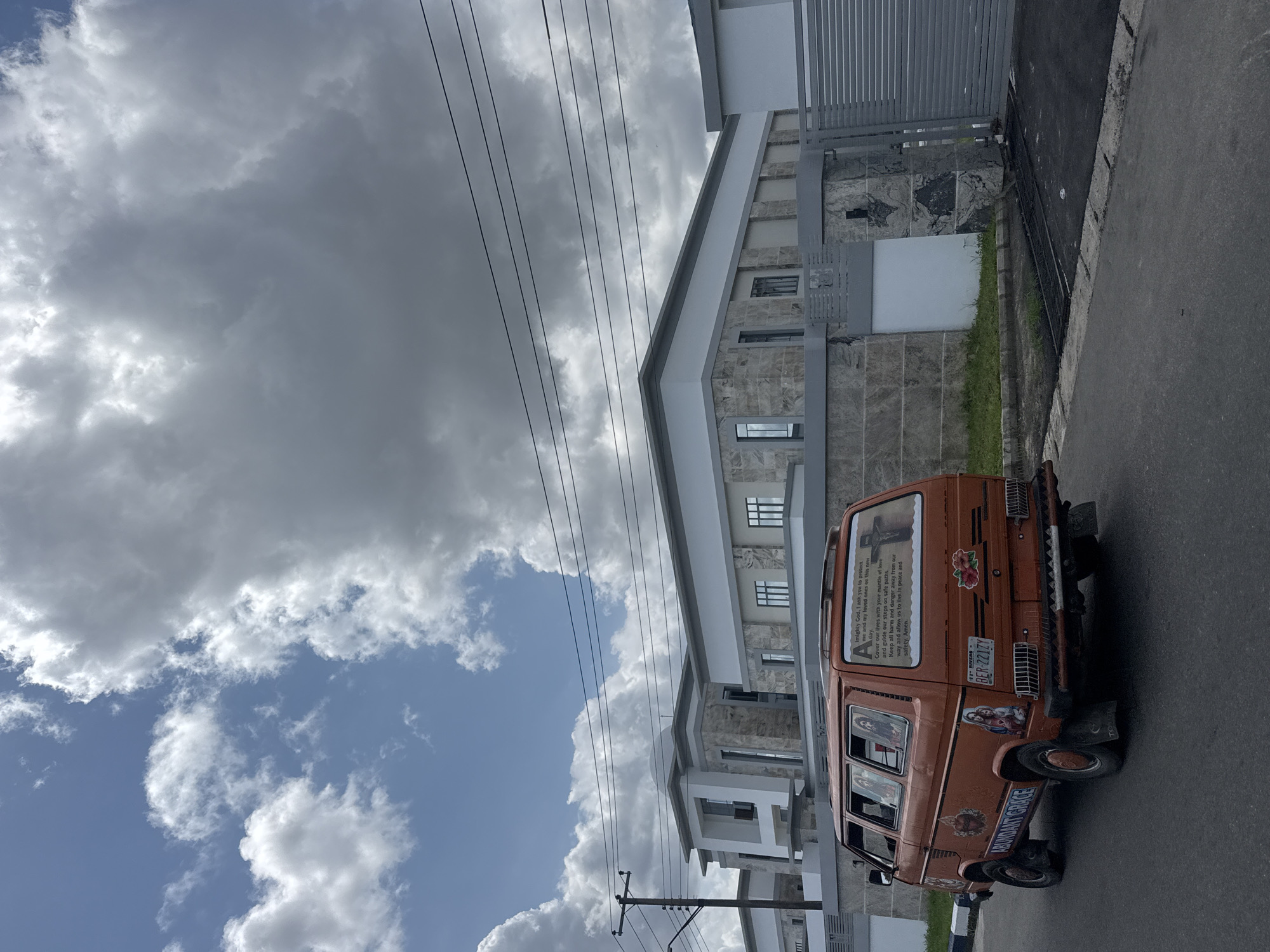 An orange van with religious iconography and decals is parked on a gray asphalt road. The license plate is visible and reads "BER-221ZY", indicating it's from Rivers State. Text on the back of the van mentions "Almighty God" and "ABUNDANT GRACE".
The van is parked next to a modern, multi-story building with gray stone accents and white trim. The building is enclosed by a gray metal fence and gate. Overhead power lines run parallel to the building. Above, a sky with puffy white and gray clouds is visible. The image is taken on a sunny day.