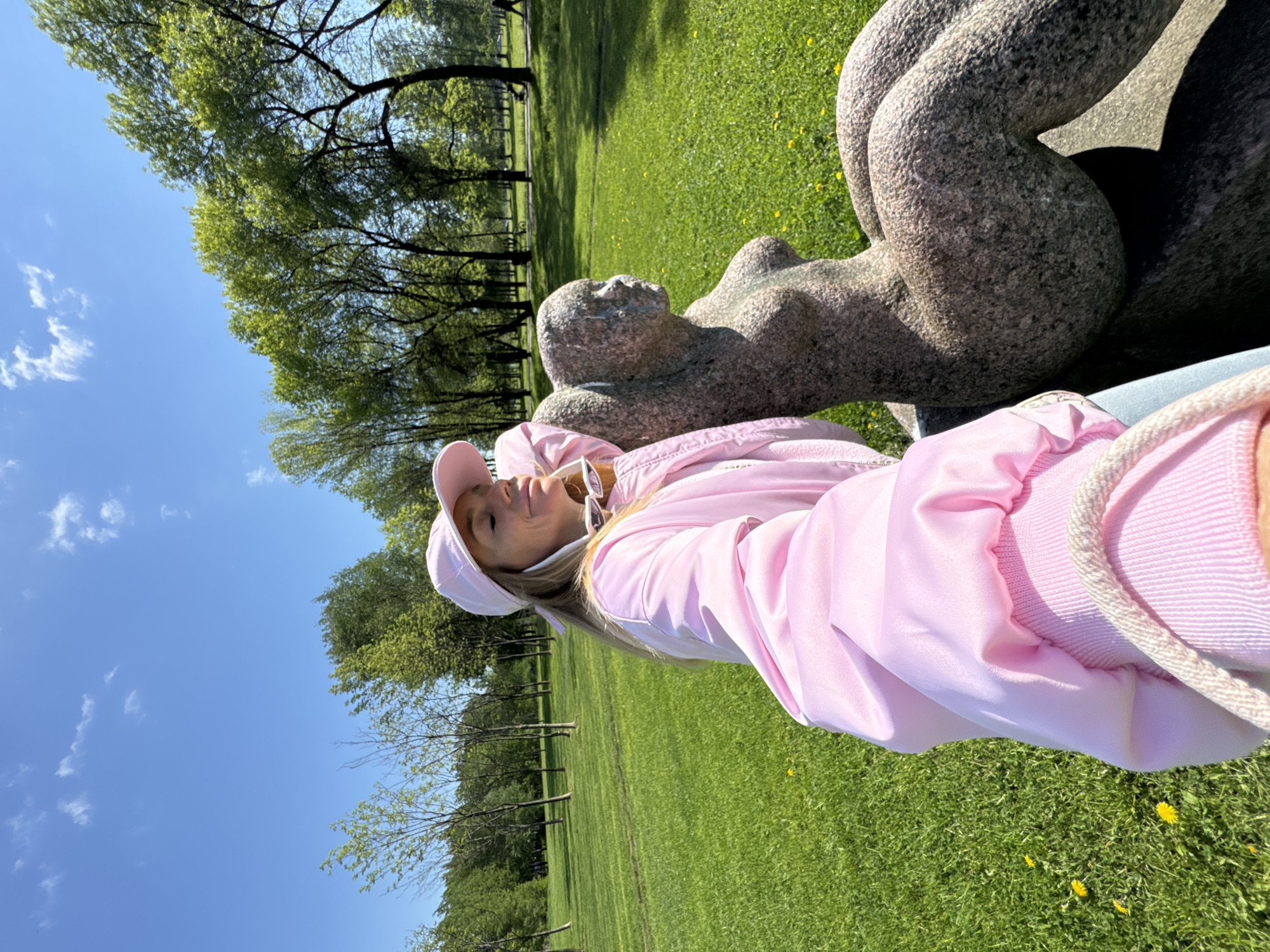 A woman in a pink tracksuit and cap lies next to a grey stone sculpture in a park. The sculpture depicts a reclining, nude human form. The park has green grass with yellow flowers, trees, and a blue sky with wispy clouds visible. The location is likely in Saint Petersburg, Russia.