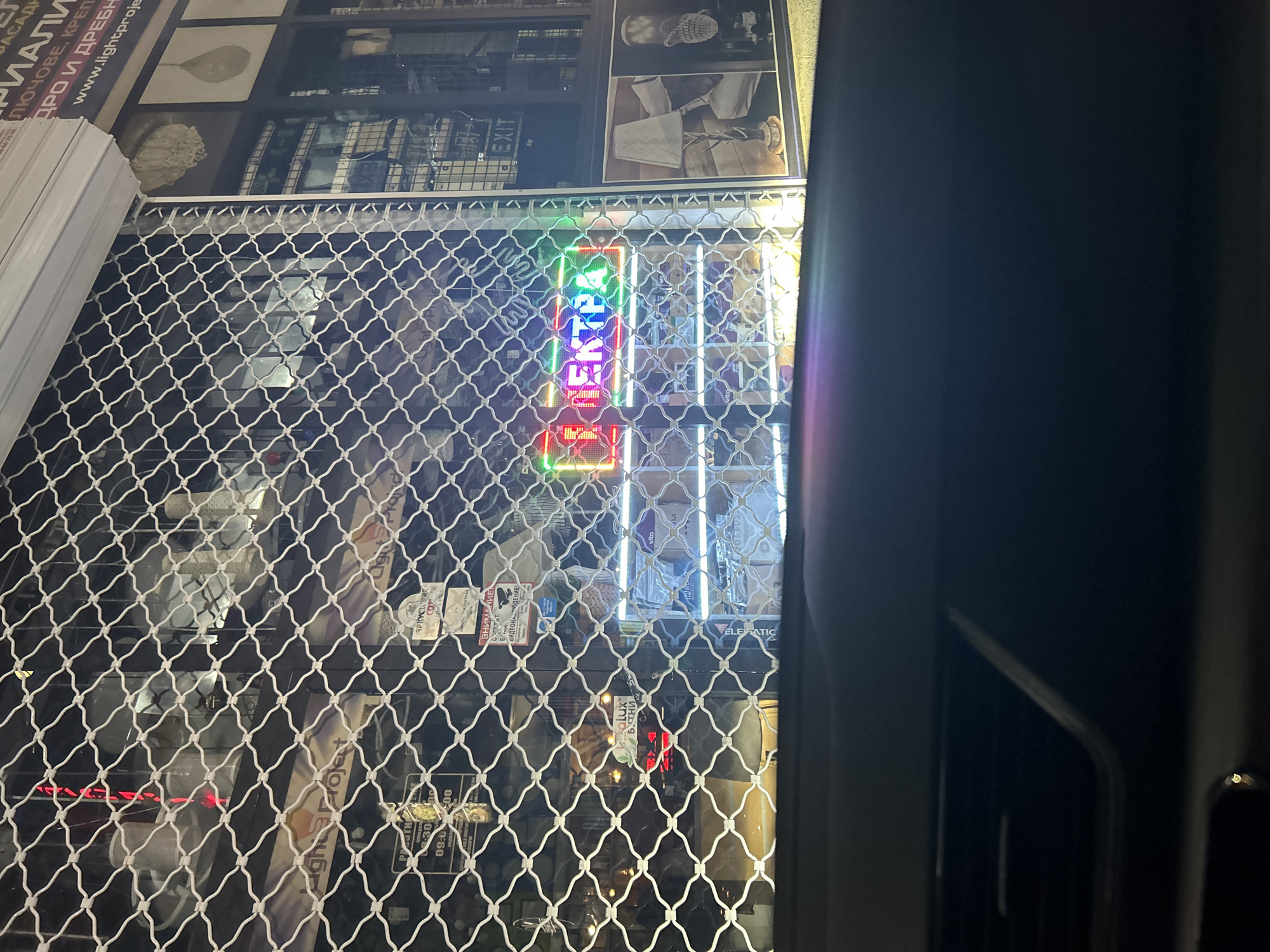 The image shows the exterior of a shop or business in Burgas, Bulgaria, viewed through a wire mesh security fence. A colorful neon sign that reads "EXTRA" is visible through the mesh. Displayed on the upper portion of the shop's exterior are a variety of images, possibly advertisements or decorative displays, set against a dark background. There are also product boxes visible. The location of the shop front appears to be within an urban area, with signs in Cyrillic, indicating the Bulgarian location. A portion of the interior is visible through the mesh and the reflections on the front glass. Part of a vehicle or a building is visible on the right side of the image.
