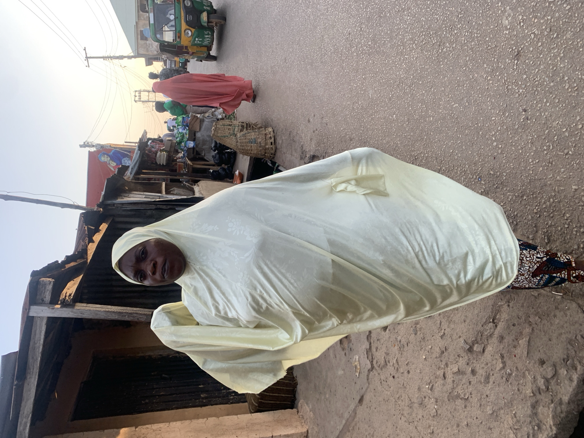 A woman is depicted wearing a light yellow full-body covering with her face visible, standing on a concrete or asphalt surface. Behind her are rudimentary buildings and a bustling street with a woman in a red robe, a tuk-tuk, and market stalls filled with various goods. The image is likely taken in Jos, Nigeria.