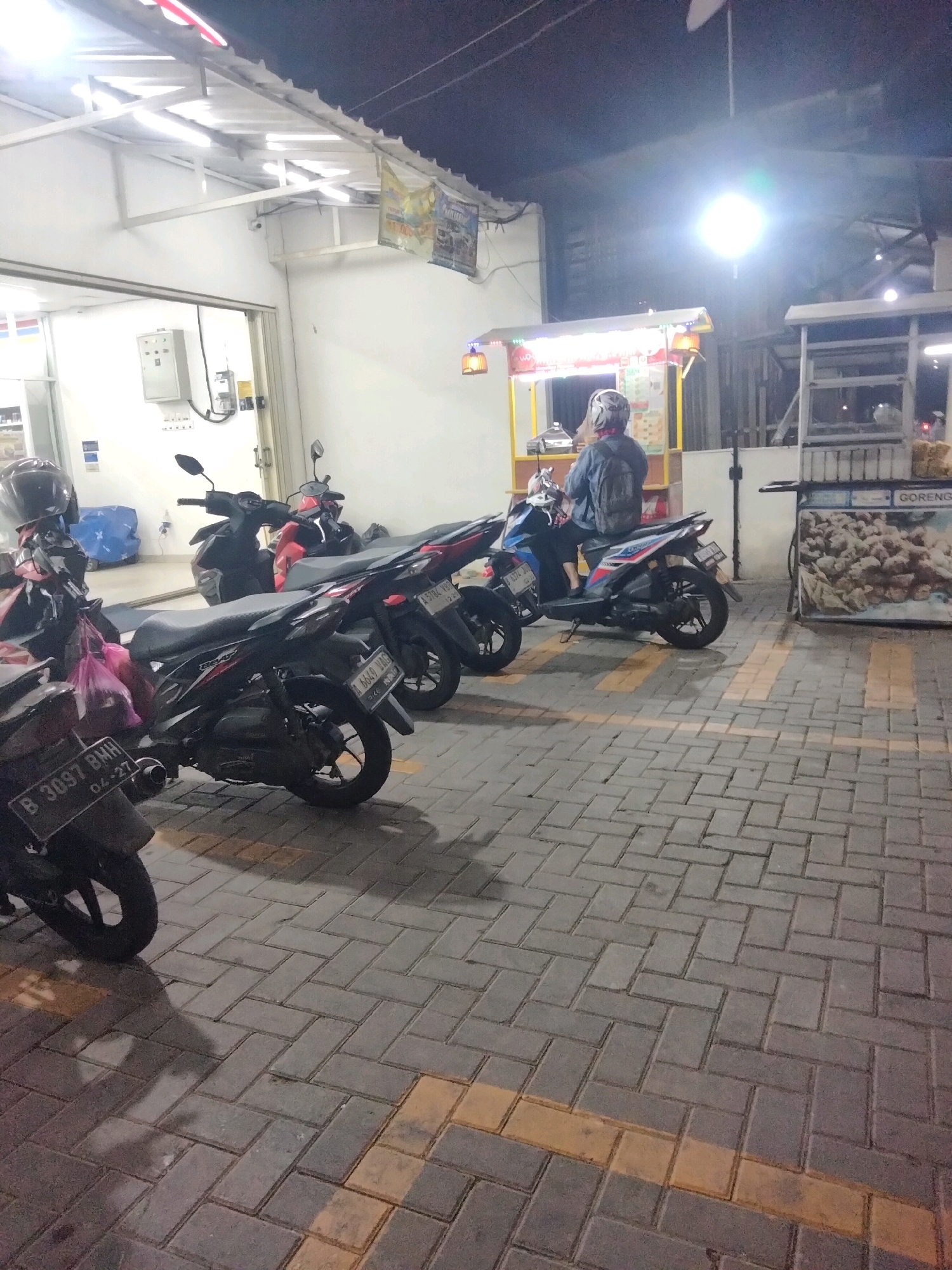 In Suka Sari, Indonesia, an evening scene unfolds outside a small establishment, likely a convenience store or eatery, illuminated by overhead lights. Paved ground, made of interlocking grey bricks with yellow painted lines, covers the area. Several parked motorcycles are visible: a black Honda Beat with license plate B 3097 BMH from 04-27 is in the foreground, to its right is another black Honda Beat with license plate B 6040 VNG. Further back, more motorcycles are parked, including a red and black one. A person wearing a helmet and a dark jacket sits on a blue and black motorcycle in front of a food stall. The stall has a yellow frame and is adorned with lights and various signage, one of which displays the word "GORENG" (fried). Another structure to the right, possibly a food cart, displays a graphic image of fried food, with "GORENG" also visible on its signage. The background shows a white building with a white roof and exposed beams. A white electrical box is mounted on the wall. The overall atmosphere is that of a typical street scene in a bustling Indonesian town at dusk or night.