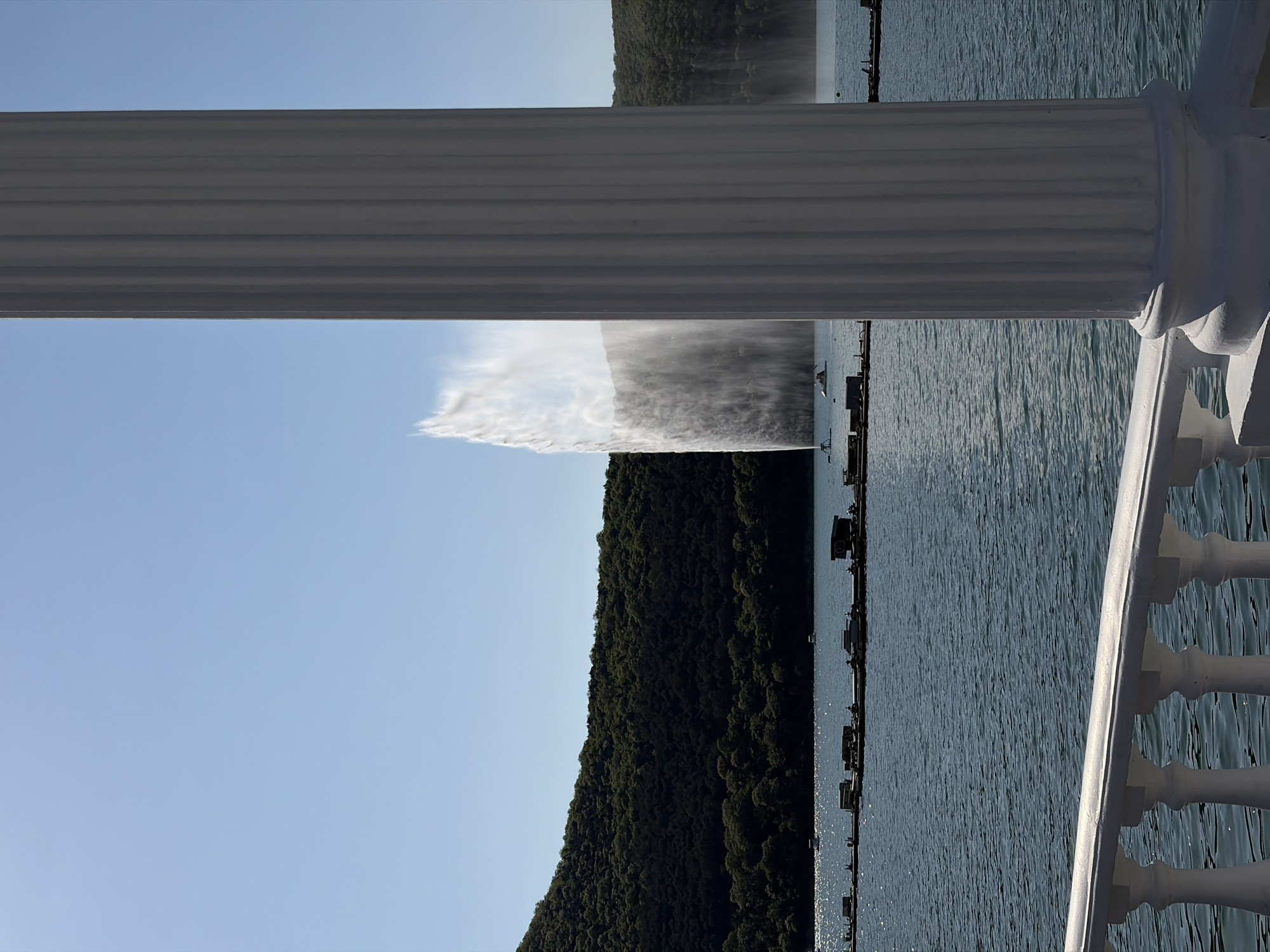 The image depicts a view from a balcony overlooking Lake Abrau in Abrau-Dyurso, Russia. A white balustrade and a fluted column frame the scene. In the distance, a large fountain sprays water high into the air, appearing as a white plume against a blue sky. A densely wooded hillside rises behind the fountain. The surface of the lake is visible, reflecting the sunlight. There appear to be docks and small structures along the shoreline. The sky is clear and blue.