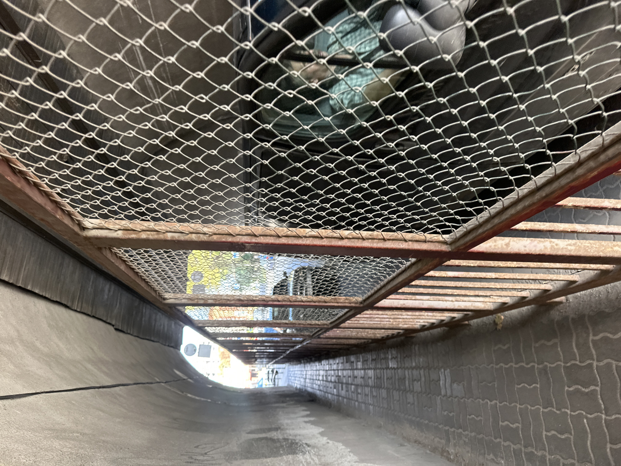 The image depicts a pedestrian walkway alongside a chain-link fence with a rusty metal frame. The walkway is paved with interlocking grey bricks and is bordered by a curved concrete wall on the left. Beyond the fence, a dark-colored vehicle is visible, with a person wearing a green plaid shirt seated in the driver's seat. The setting appears to be a tunnel or an underpass in the city of Ternopil, Ukraine, indicated by the architectural style of the walkway and the presence of a street sign visible in the distance, which reads "ВУЛ. БАНДЕРИ" (Bandery Street). The overall scene is dimly lit, with natural light filtering in from the opening of the tunnel at the far end.