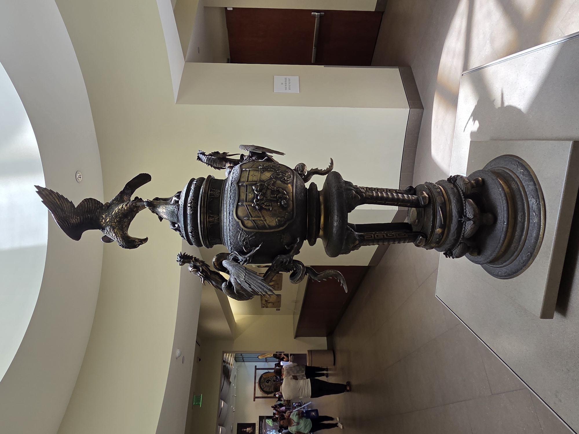 An ornate, dark-colored bronze incense burner with intricate dragon and bird motifs stands on a pedestal in the center of a museum hall. The burner is topped with a detailed sculpture of an eagle. Sunlight streams through windows, casting shadows on the polished floor. In the background, visitors stroll through the museum, with some artwork visible on the walls and a doorway leading to another section. This scene is located in the Bowers Museum in Santa Ana, California, United States of America.