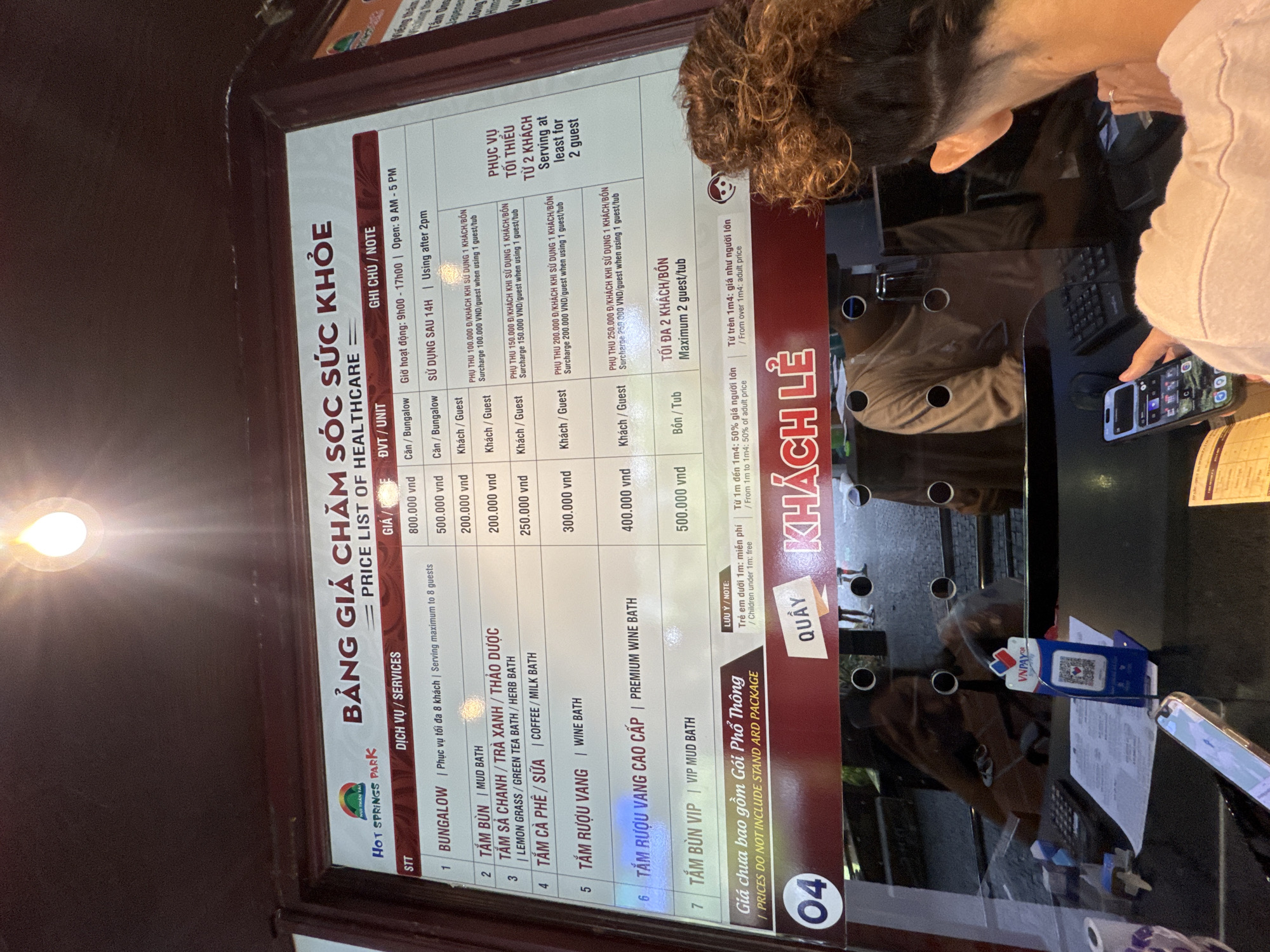 The image displays an indoor scene at a service counter, likely within the "Hot Springs Park" in Đà Nẵng, Vietnam. A large, angled price list board for "BẢNG GIÁ CHĂM SÓC SỨC KHỎE" (PRICE LIST OF HEALTHCARE) dominates the foreground, detailing various services and their costs in Vietnamese Dong (VND).
The services listed include Bungalow access (800,000 VND for up to 8 guests), Mud Bath (ranging from 200,000 VND to 500,000 VND), Lemon Grass/Green Tea/Herb Bath, Coffee/Milk Bath, Wine Bath, Premium Wine Bath, and VIP Mud Bath. Operating hours are noted as 9:00 AM - 5:00 PM, with specific notes about using services after 2:00 PM. The board also outlines surcharges for single guests (ranging from 100,000 VND to 250,000 VND) and details for "KHÁCH LẺ" (Individual Guests), including pricing for children (free under 1m, 50% of adult price for 1m-1m4, and full adult price for over 1m4). A disclaimer states that prices do not include the "STANDARD PACKAGE."
On the right, a person with curly brown hair is partially visible, leaning over a counter, holding a smartphone. A VNPAY QR code stand is present on the counter, indicating available mobile payment options. The word "QUẦY" (Counter) is seen on the side of the price board. The scene suggests a customer is at a reception or booking counter, possibly in the process of purchasing or inquiring about services.