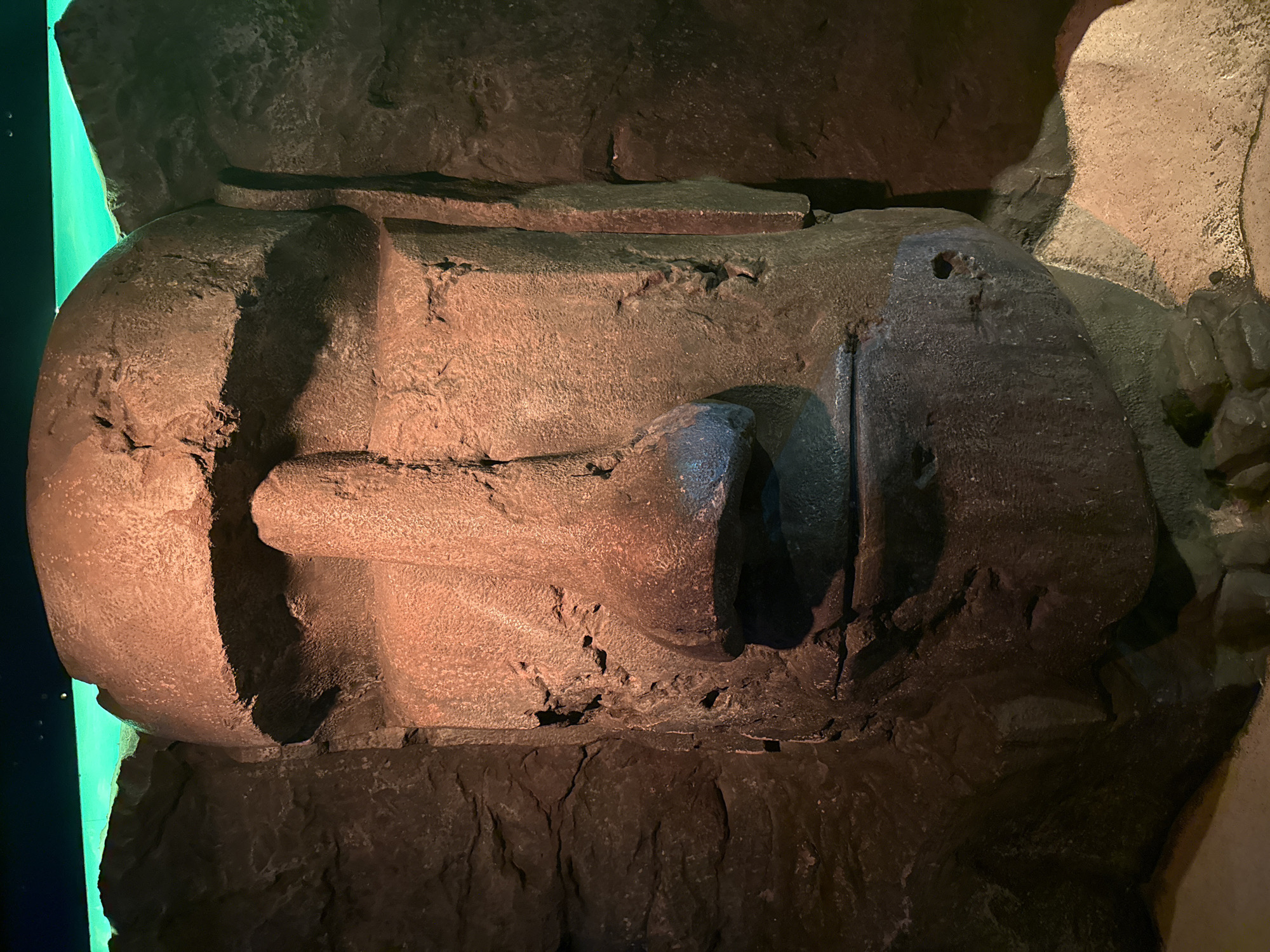 The image shows a monumental sculpted head, possibly of Ramesses II, resting on a dark, uneven stone base. The head, rendered in dark-brown stone, is depicted in profile. Details include a prominent nose, a suggestion of an eye, and a stylized beard. The head is illuminated by a warm, reddish light. To the left is a panel with a bright green glow. The surrounding environment is dark and rocky. This sculpture is located at The British Museum in London, United Kingdom.