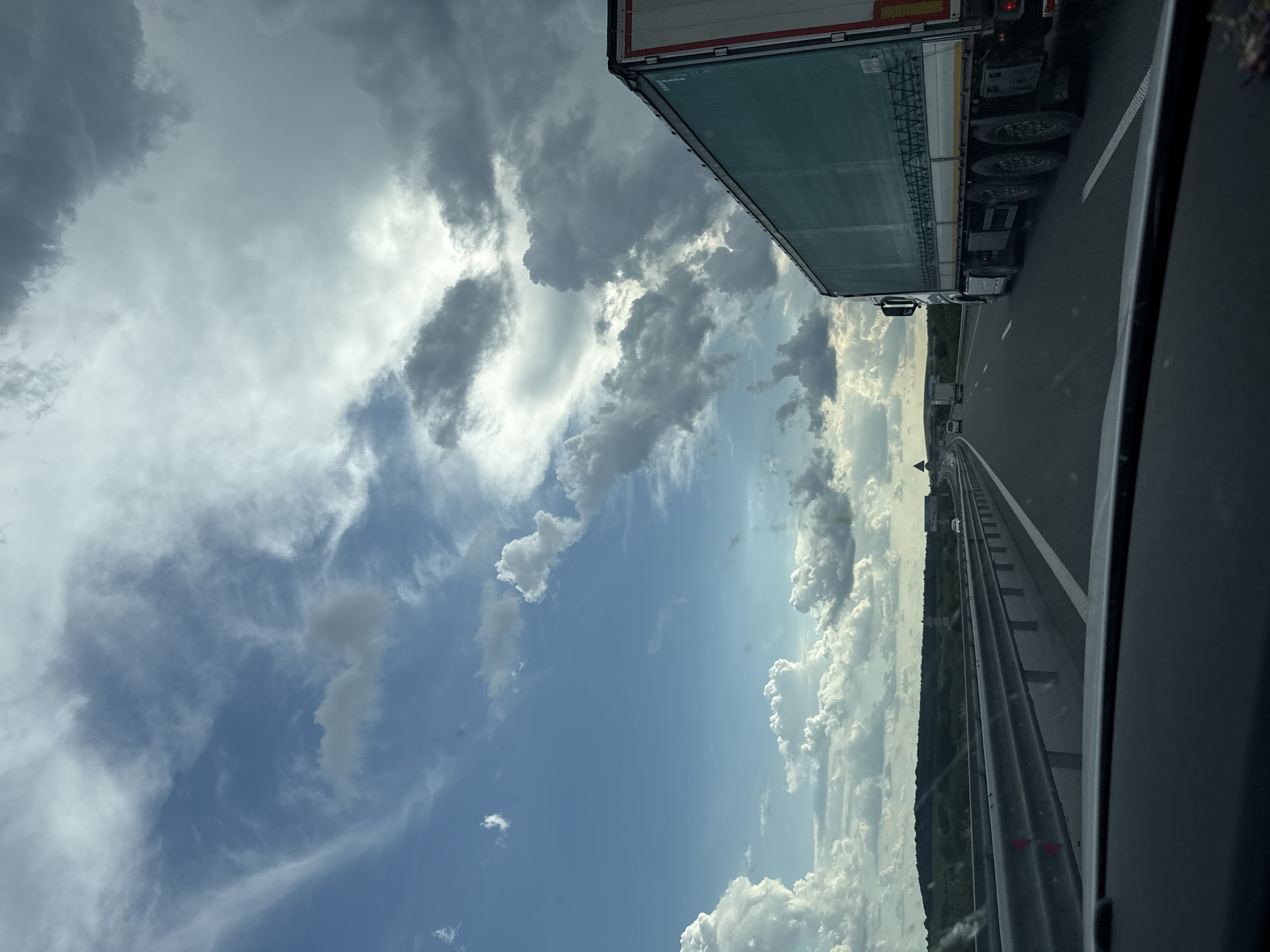 An overcast sky with scattered clouds is visible above a highway with a large semi-truck in the right lane. The highway is lined with a metal guardrail and has white lane markings. In the distance, rolling hills covered in vegetation can be seen. The image appears to have been taken from the perspective of a passenger in a vehicle traveling on the highway. There are no people or buildings visible in the frame. The location is described as Yazdach, Bulgaria.