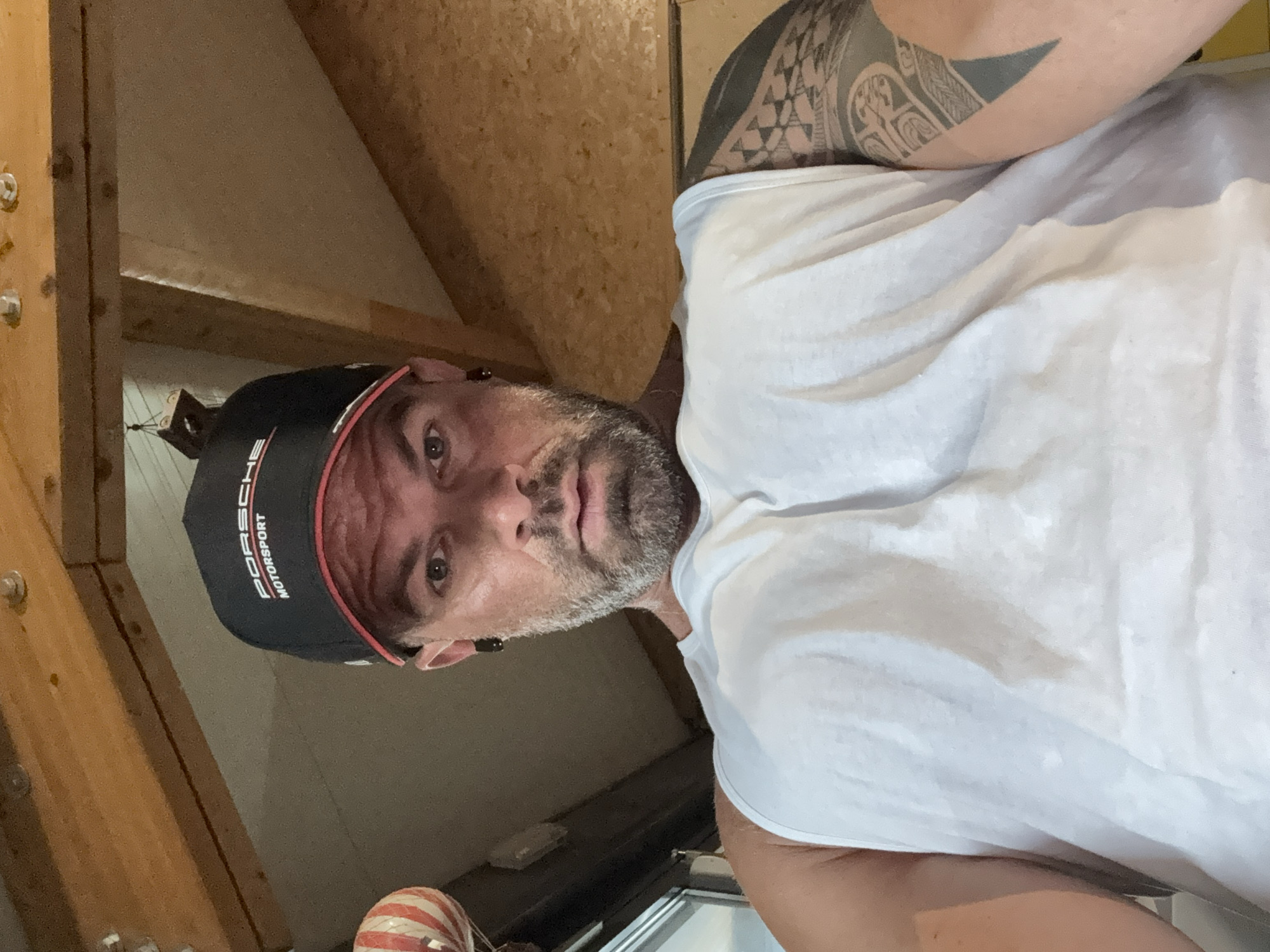 A man wearing a white tank top and a black Porsche Motorsport cap is looking at the camera. The man has a gray beard and a tattoo on his left arm. The location appears to be an attic, as indicated by the wooden beams and plywood in the background. The location is not identifiable as Jemelle, Belgium based on visual cues.